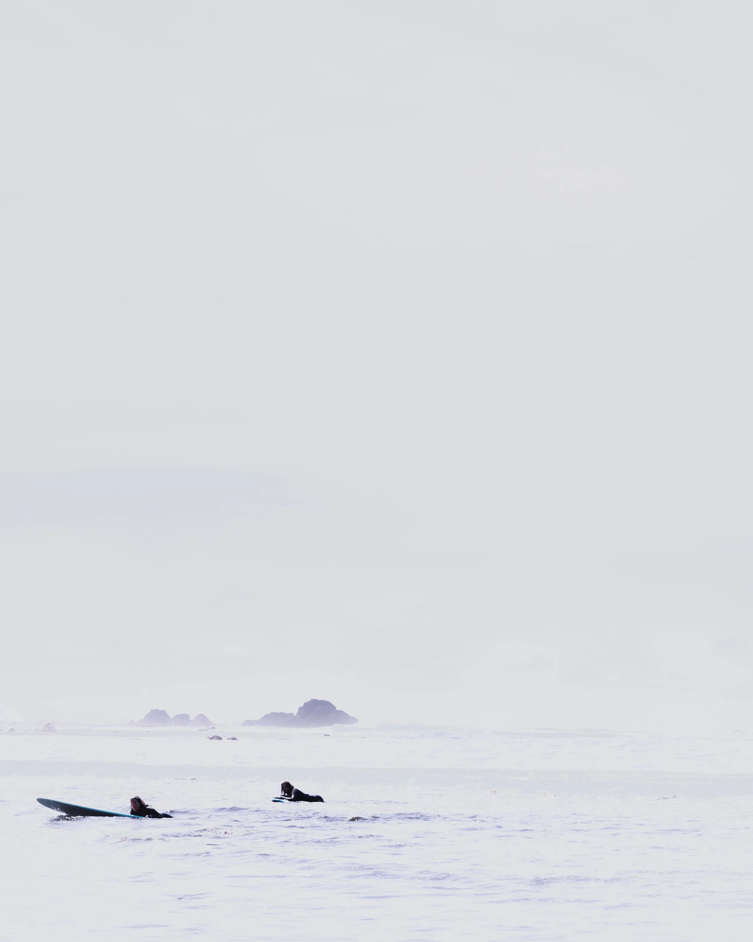 Two surfers lying on surfboards in the ocean with waves, ocean rocks in the distance, overcast sky, and distant rocky islands on the horizon.