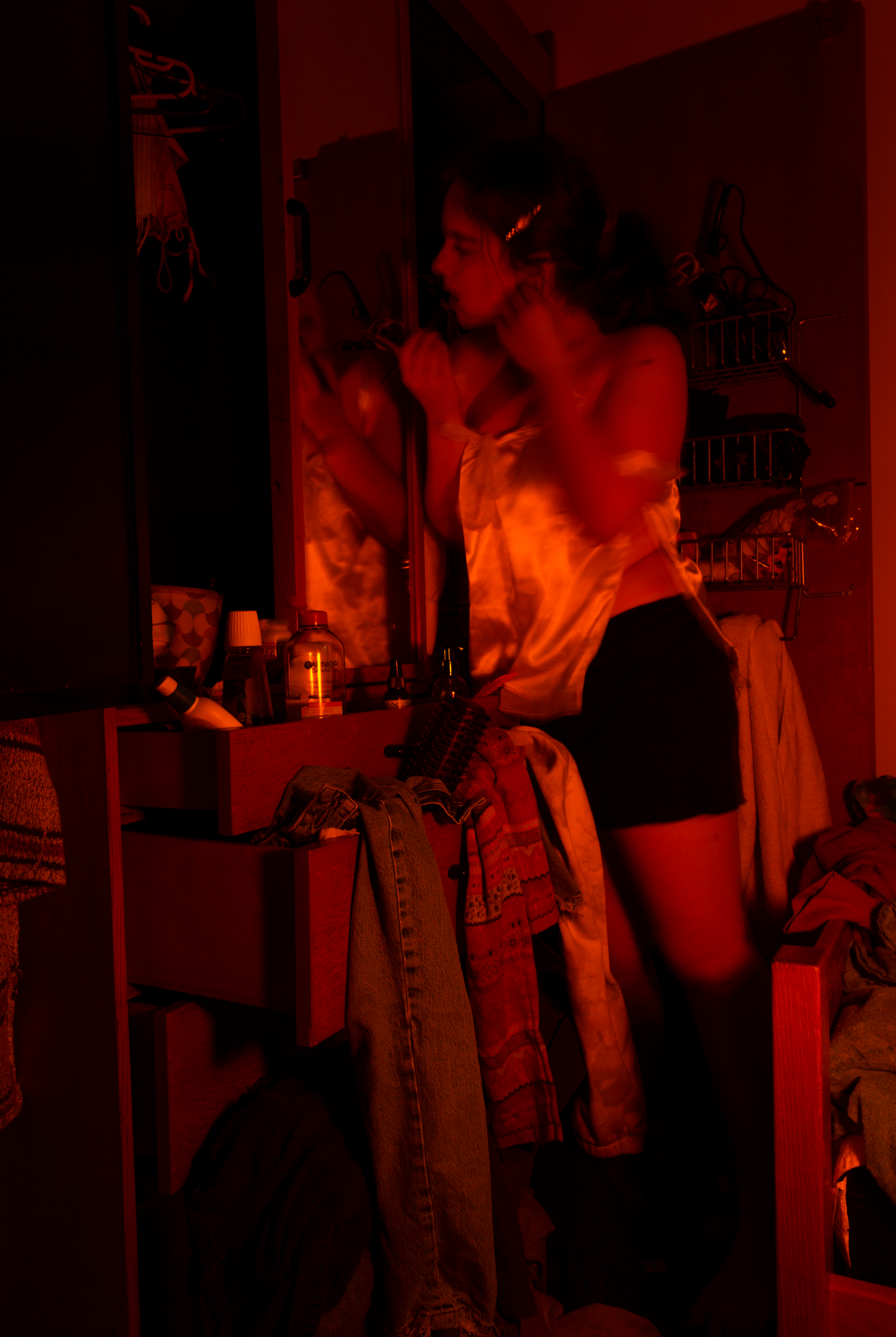 A woman with dark hair tied with a hair clip is looking into a mirror while adjusting her earrings in a dimly lit room bathed in red light. The room appears to be a closet or dressing area with shelves containing toiletries, clothes, and other person