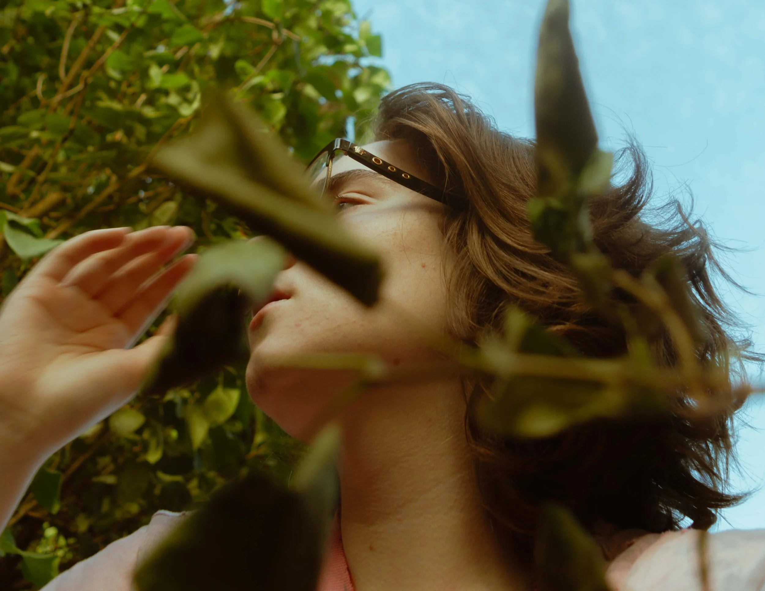 A person with curly brown hair and glasses examining a leafy plant outdoors against a blue sky.