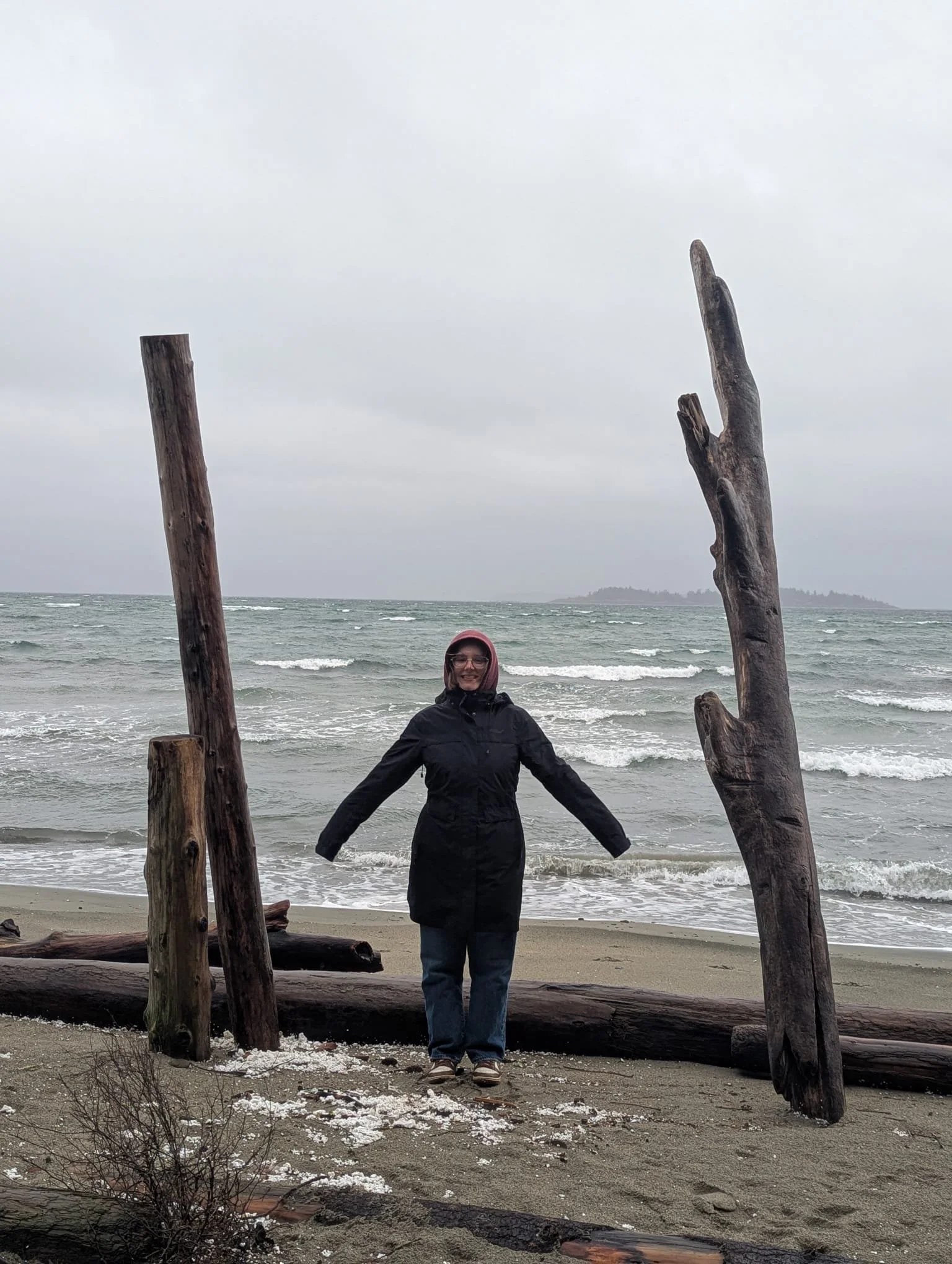 A woman wearing glasses, a black jacket, and jeans, standing on a beach with ocean waves in the background. She is smiling with arms outstretched between large driftwood logs and weathered wooden posts. The sky is overcast.