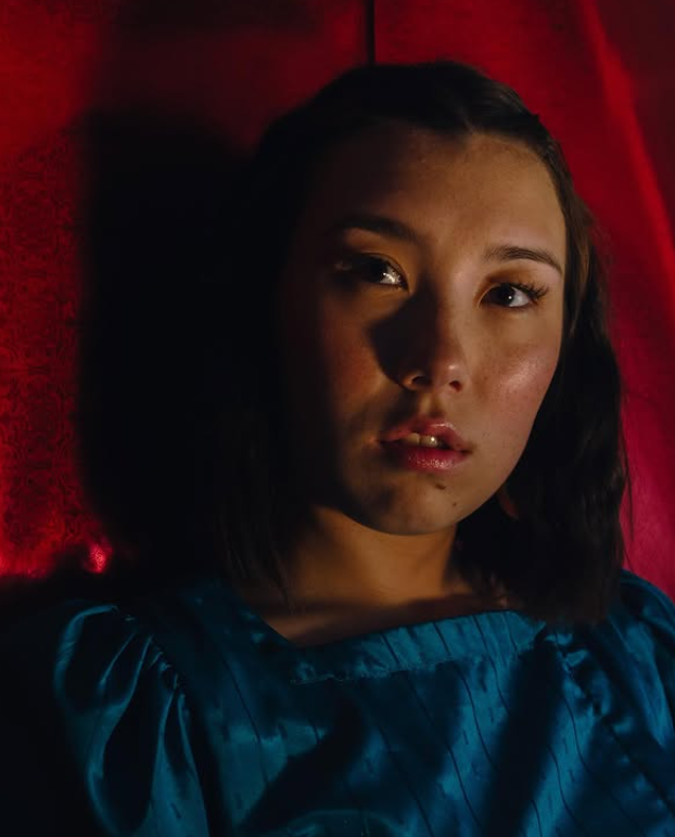 A young woman with dark hair and fair skin lying on a red surface, wearing a blue top, with a serious expression.