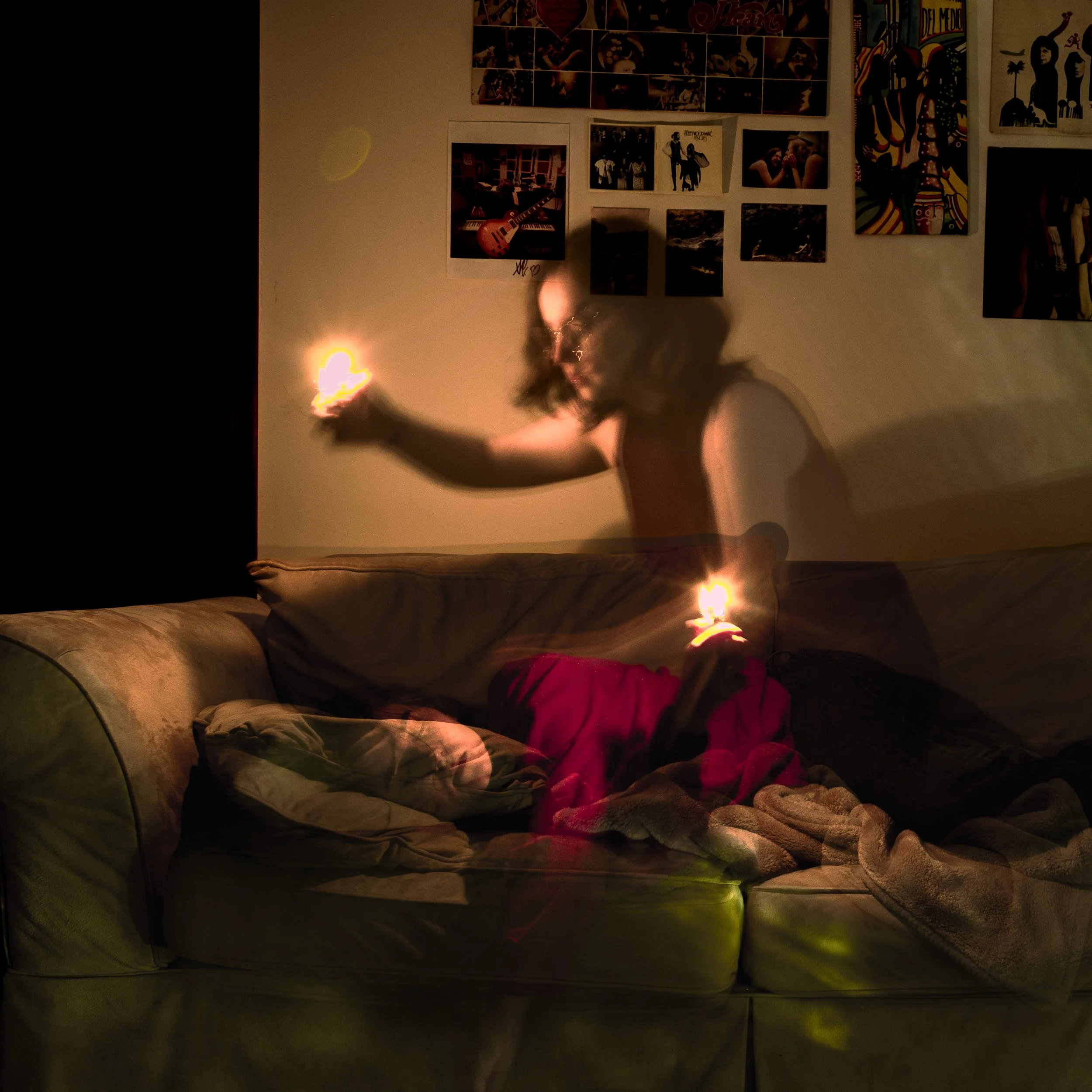 Dimly lit room with a wall decorated with numerous posters and pictures, a person with curly hair holding a lighter or match in the foreground, and a sofa with cushions at the bottom of the image.
