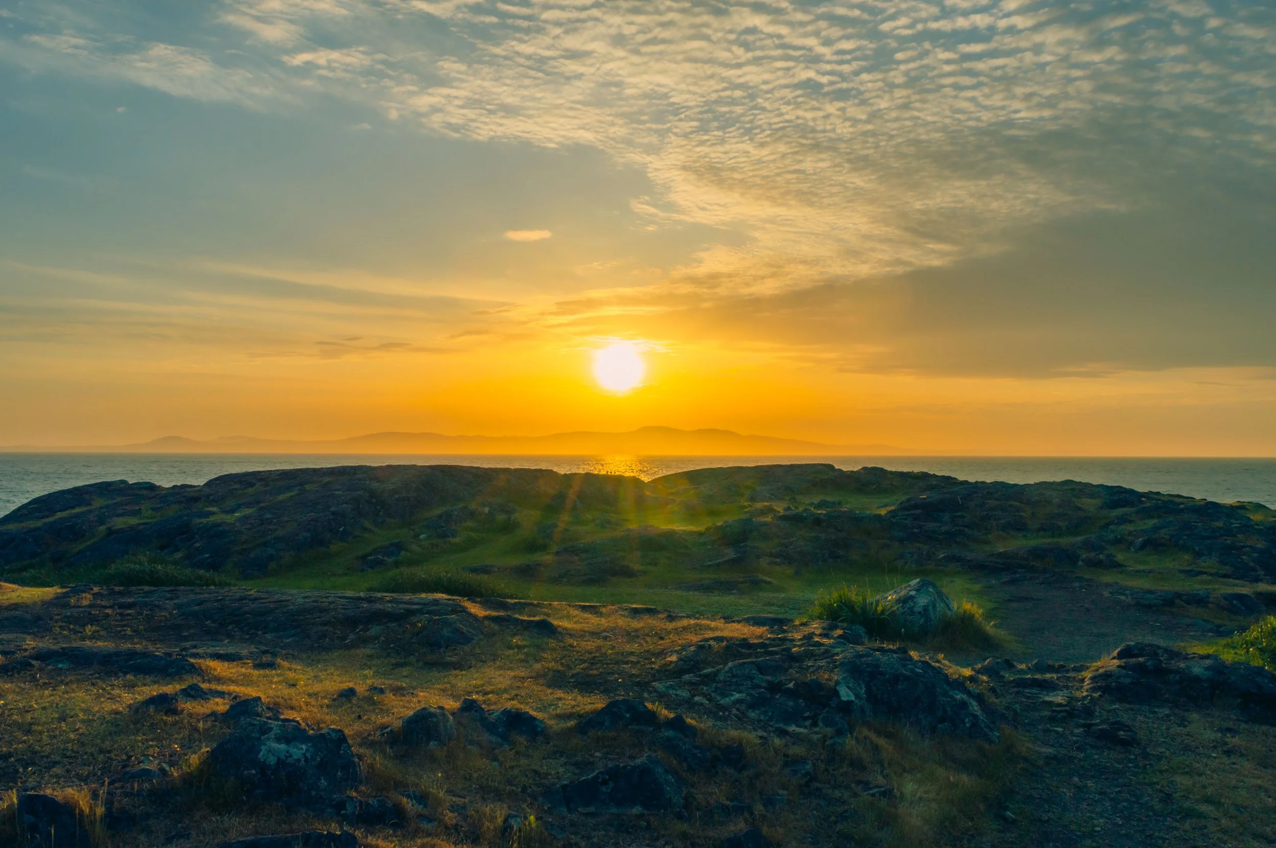 Sunset over rocky coastline with grassy landscape in foreground and distant mountains.