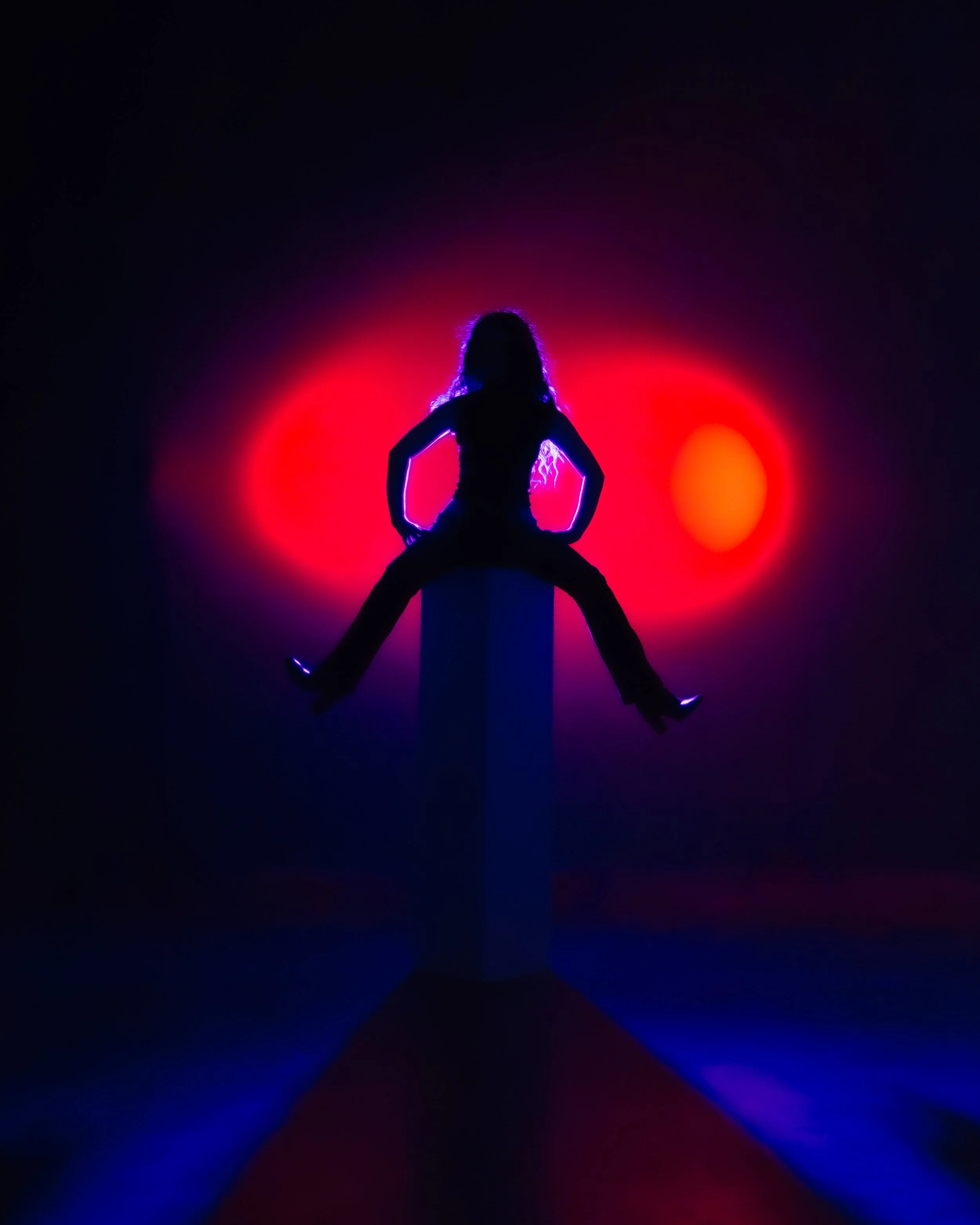 Silhouette of a woman with curly hair, sitting in a kneeling pose with hands on her hips, against a background of colorful red, pink, orange, and blue lights creating a glow effect.