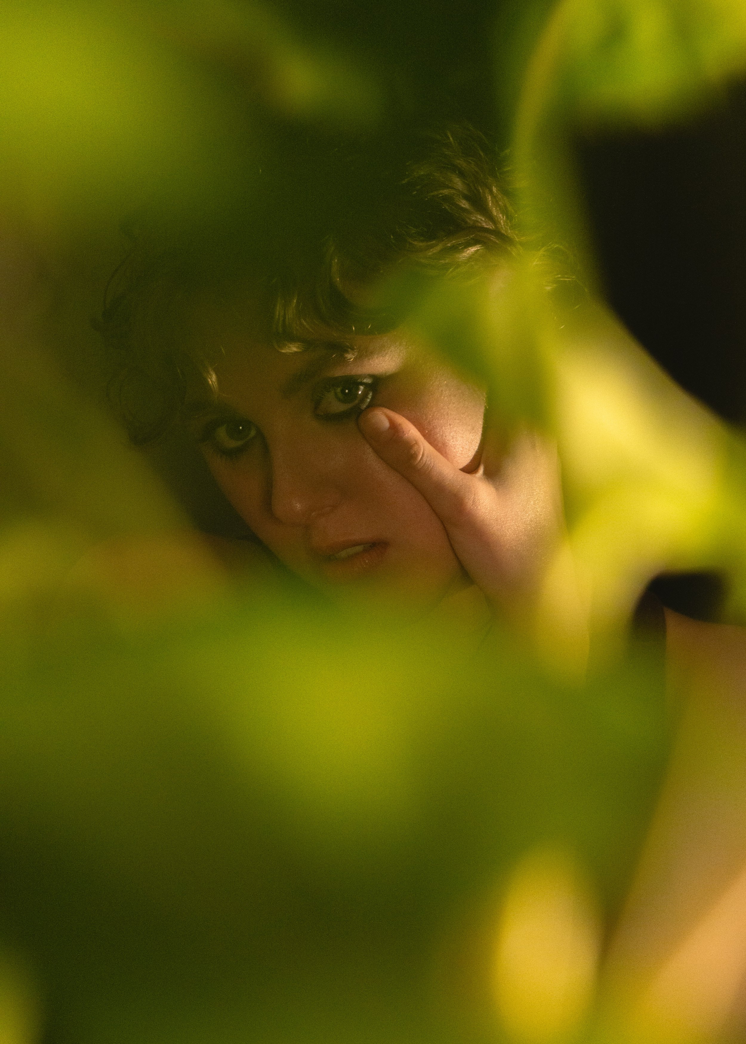 Close-up of a woman with curly hair, covering part of her face with her hand, looking through a gap in green plants.