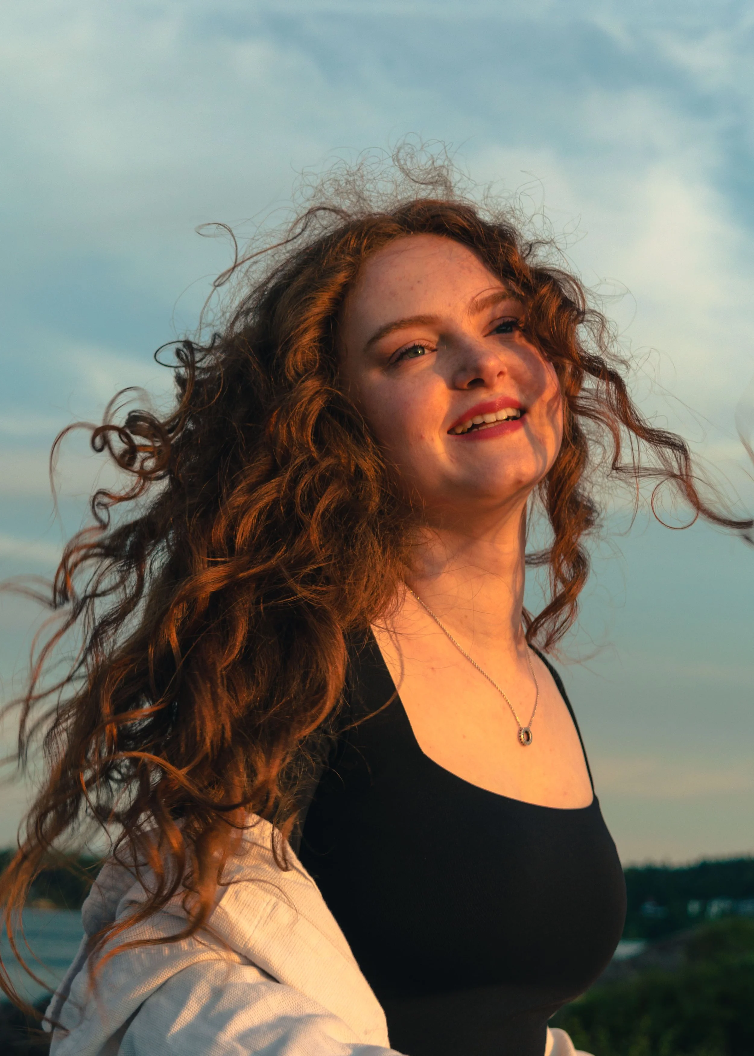 A woman with curly red hair and freckles smiling outdoors at sunset, wearing a black top and beige jacket.
