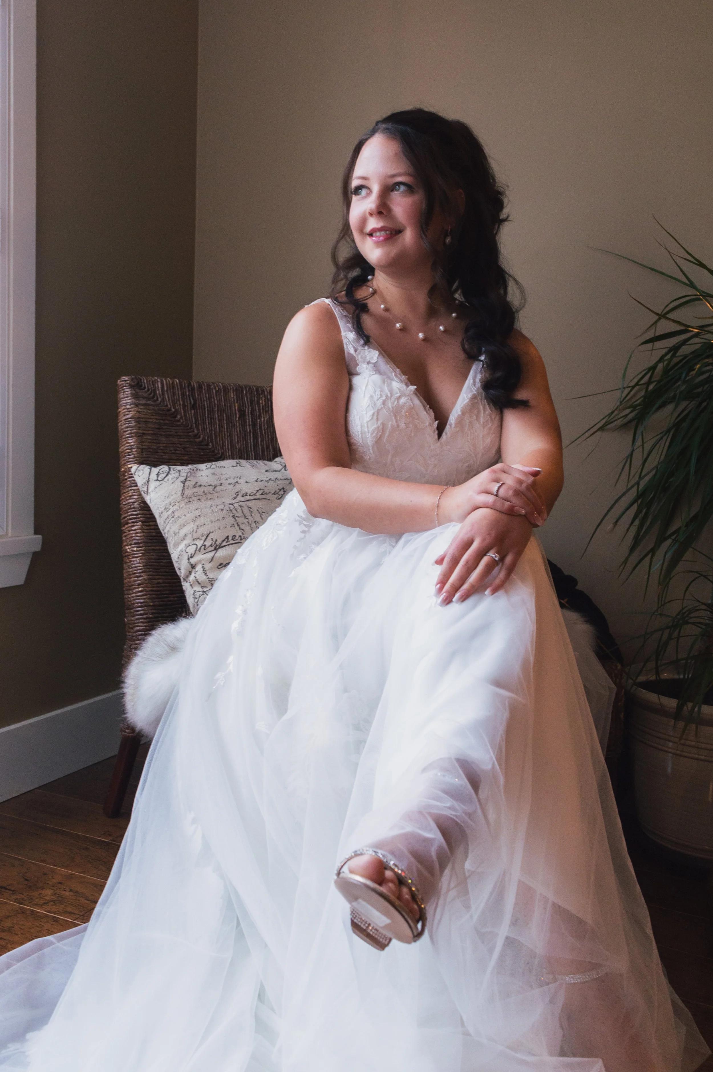 A woman in a white wedding dress sitting on a chair, with dark hair styled in loose curls, wearing a pearl necklace and jewelry, smiling and looking to the side in a softly lit room.