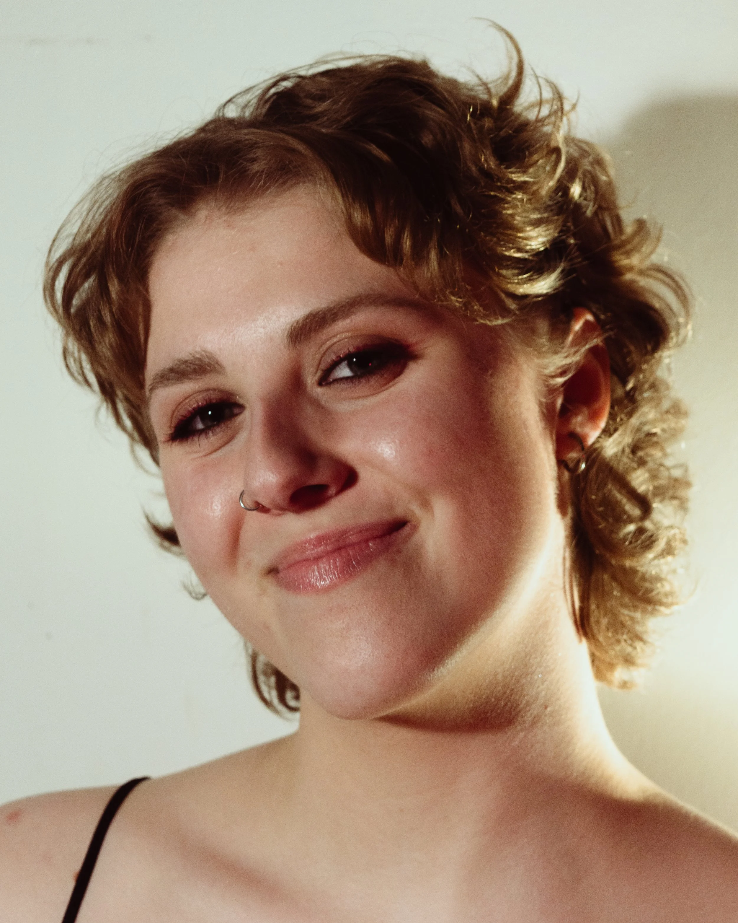 Close-up of a smiling woman with curly, short red hair and a septum piercing, wearing a black spaghetti strap top, against a plain light-colored background.