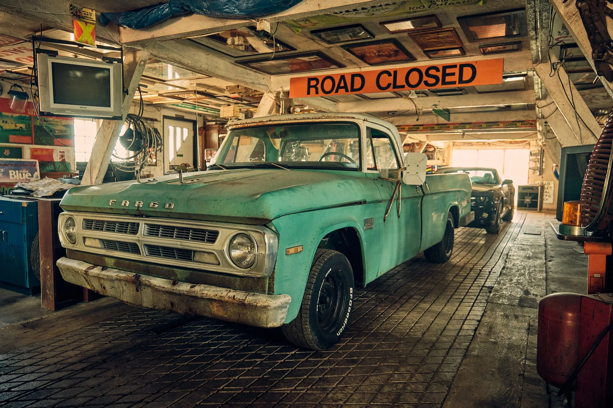 Vintage garage interior with an old faded green pickup truck inside. The garage is cluttered with tools, posters, and miscellaneous items. A bright orange sign reads 'ROAD CLOSED.'