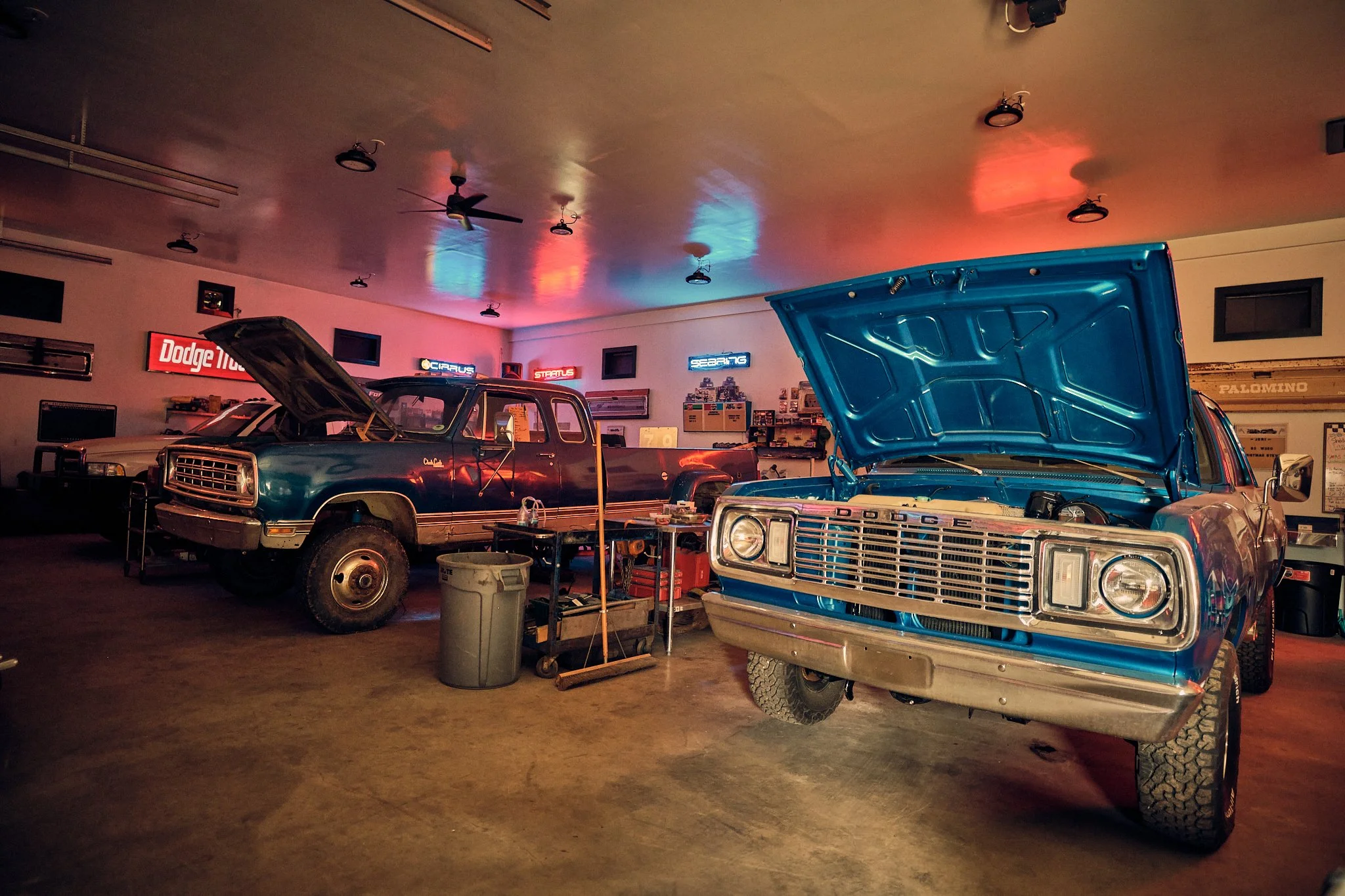 A garage or workshop with classic cars, some with open hoods, illuminated by neon signs on the wall.