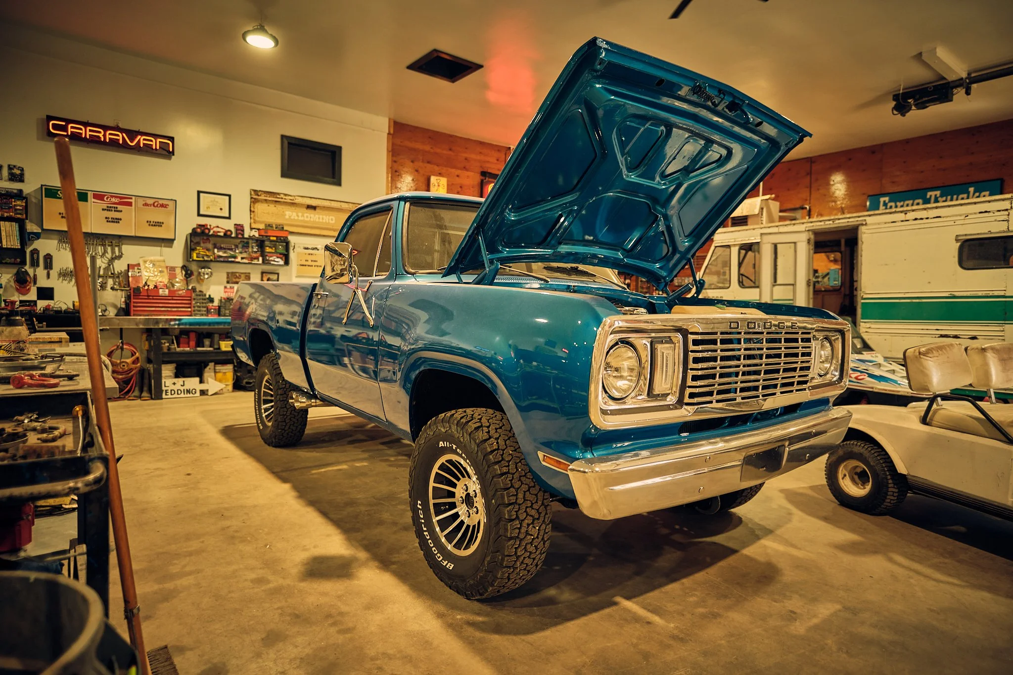 A vintage blue Dodge pickup truck with its hood open, parked inside a garage filled with tools and memorabilia.