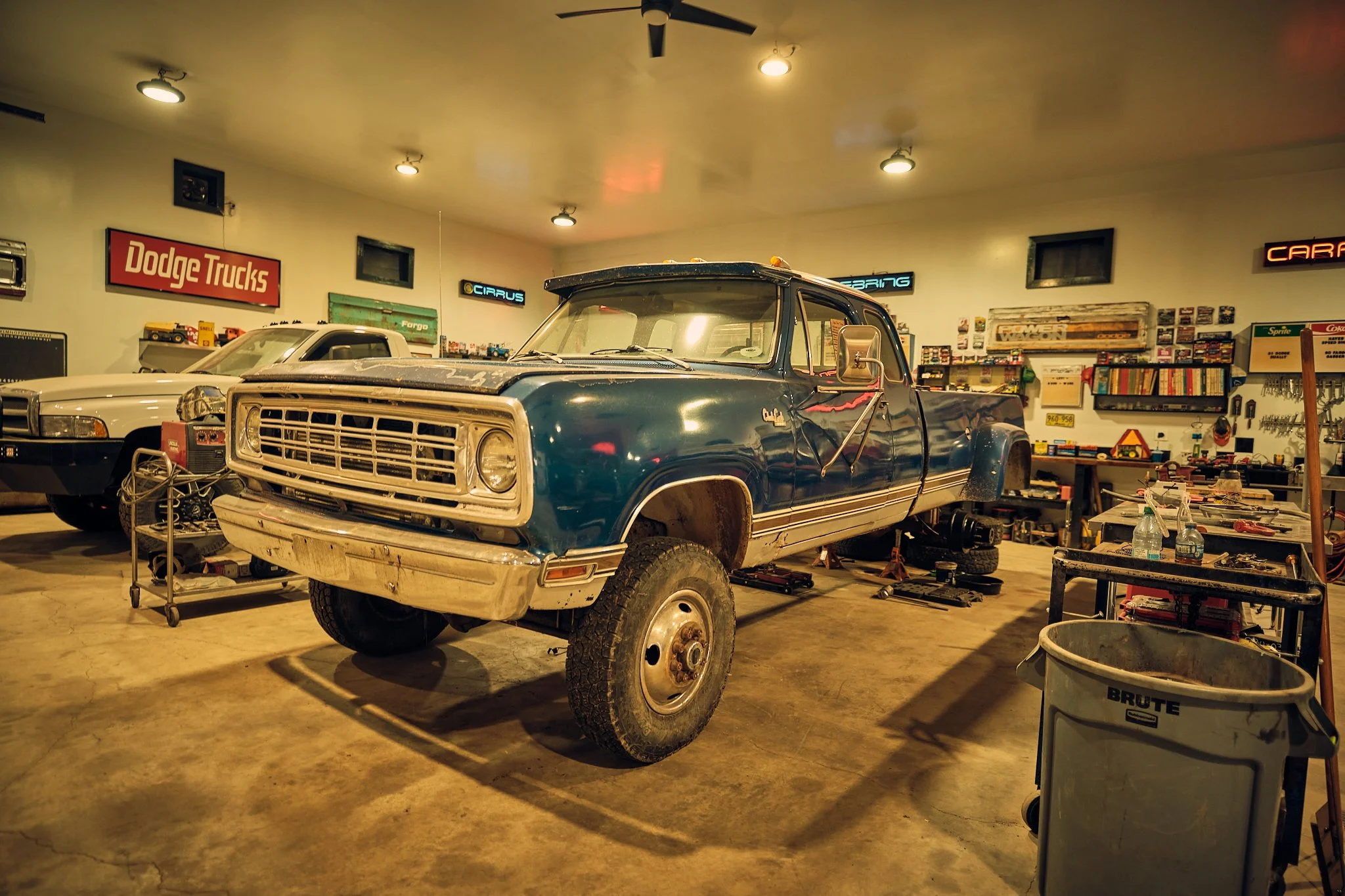 A vintage blue pickup truck in a garage workshop with various tools and signs on the walls, including a Dodge Trucks sign.