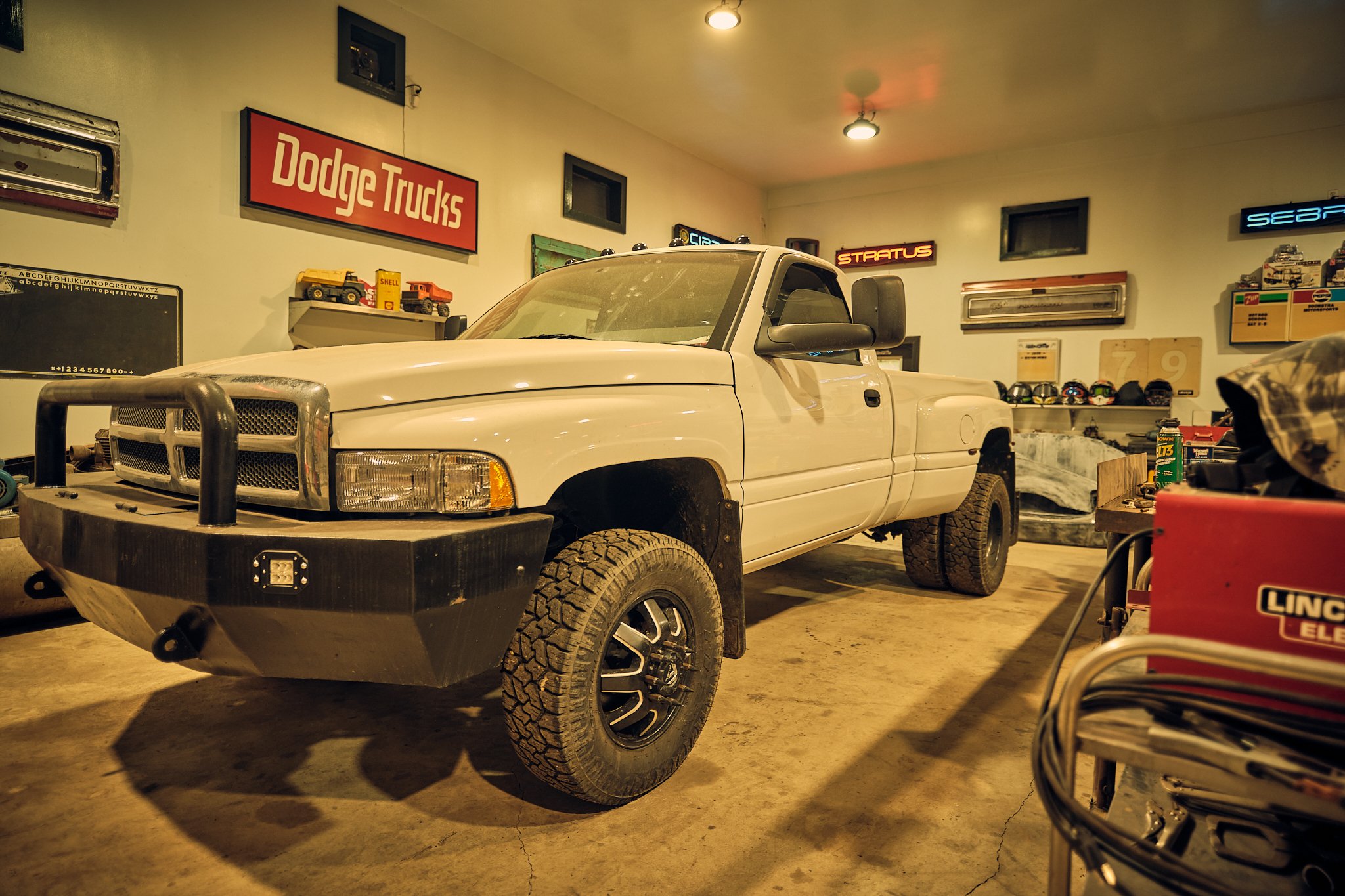 A white pickup truck is parked inside a garage or workshop, surrounded by various tools, signs, and collectibles related to trucks and automotive culture.