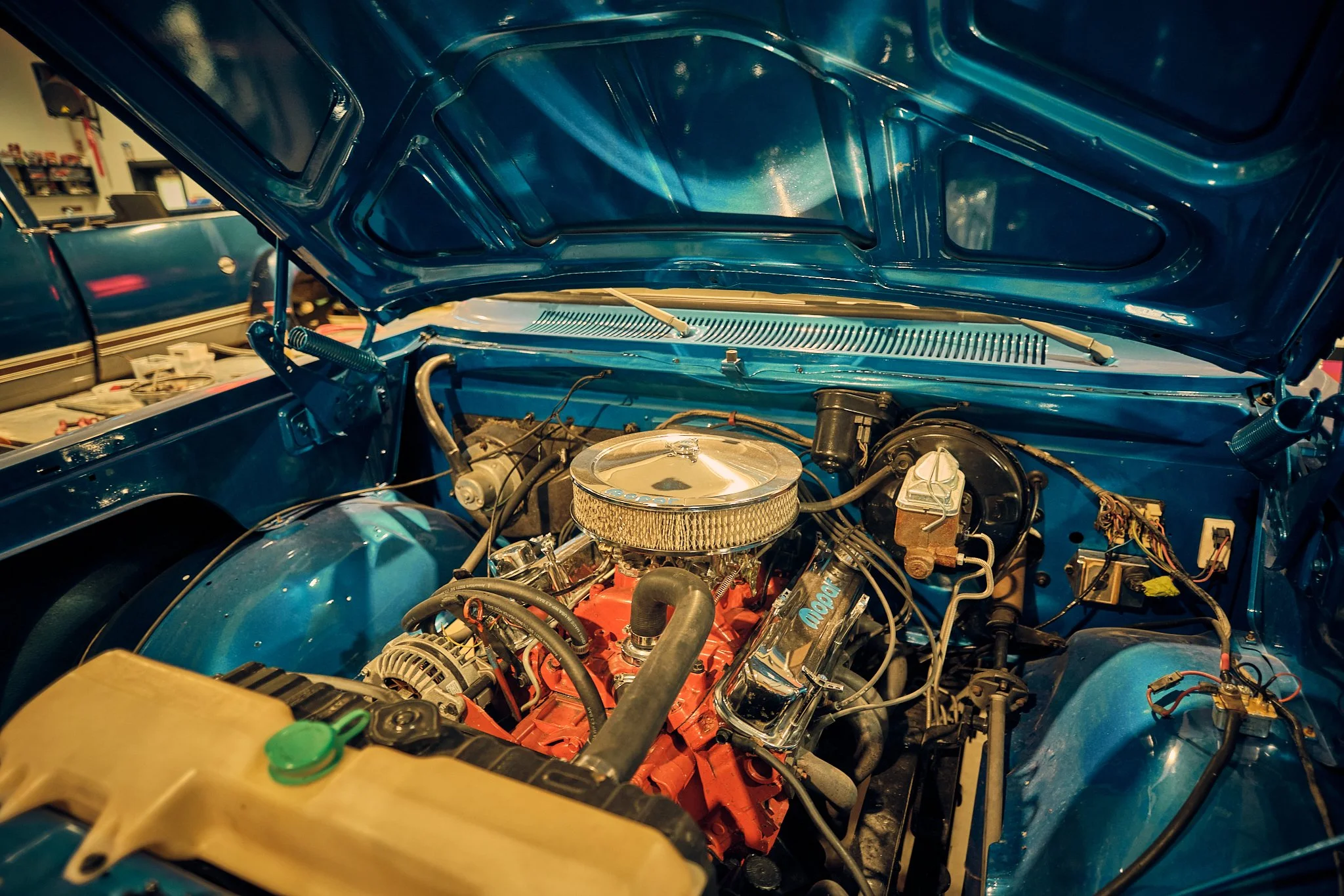 View of a car's engine bay with the hood open, showing a V8 engine with a chrome air cleaner, surrounded by various hoses and wires.