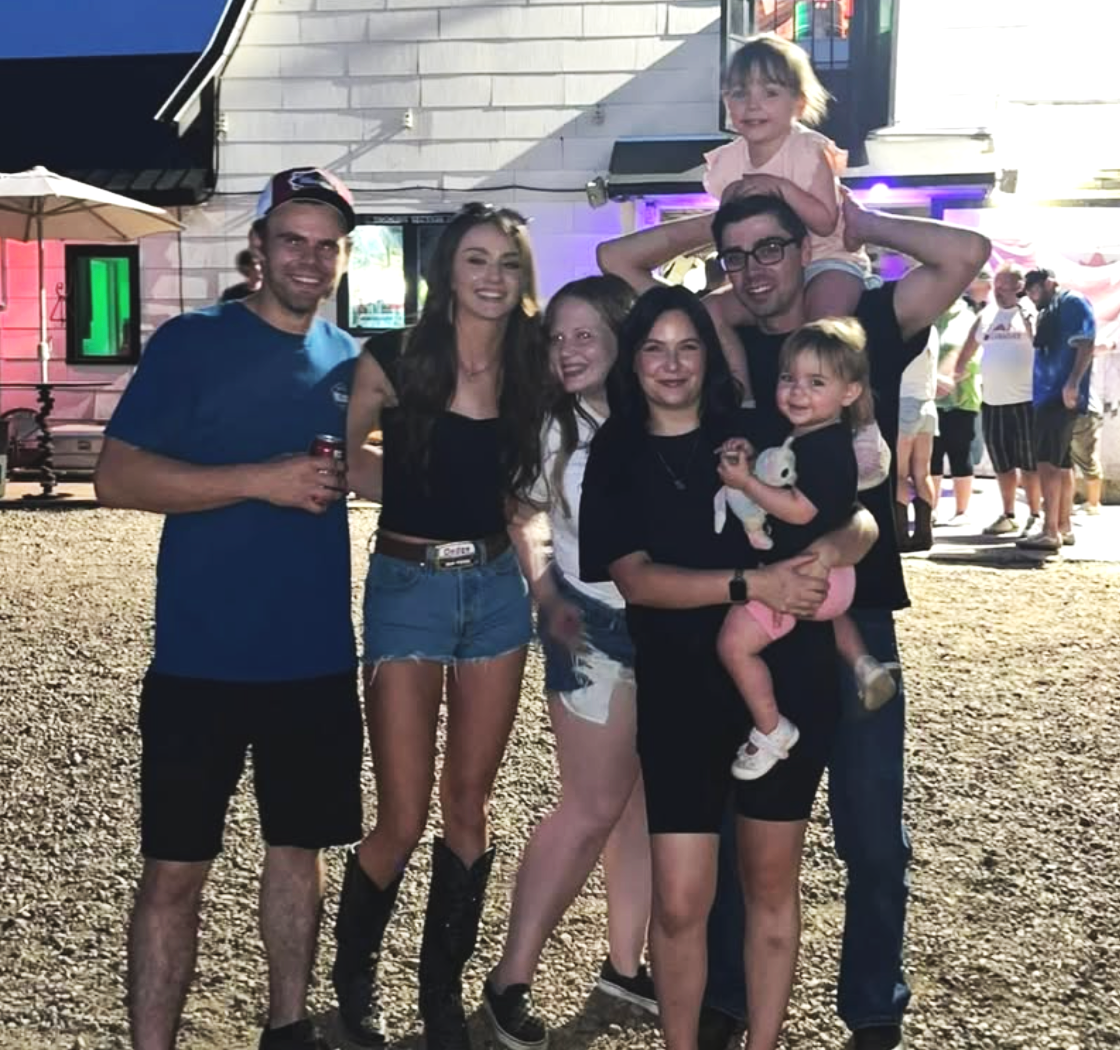Group of six people, including two children, posing together at an outdoor event during evening time, with a white building and festival lights in the background.