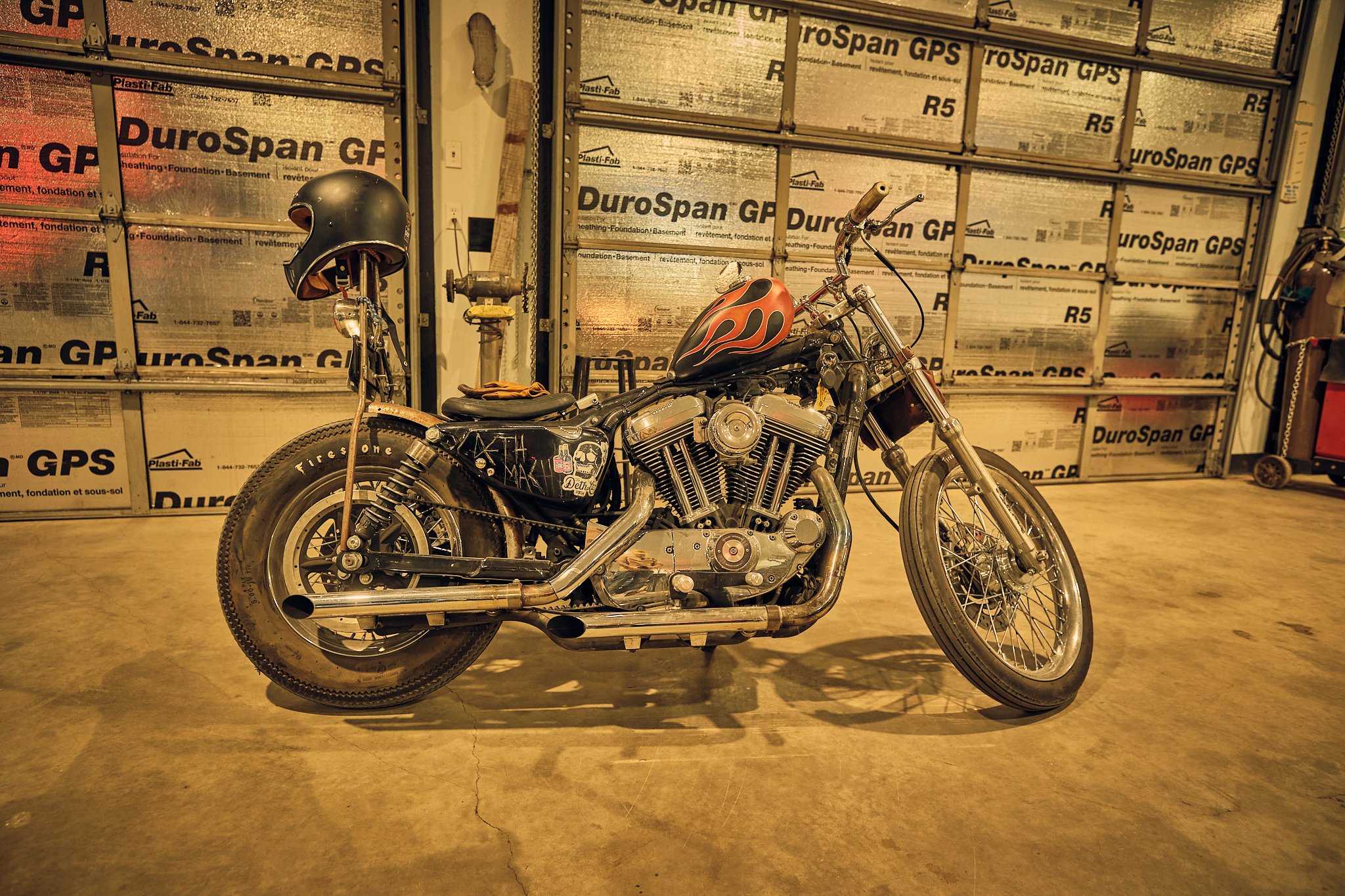 A black motorcycle with flame decals on the gas tank, parked inside a garage with unfinished walls covered in insulation labeled 'DuroSpan GP.'