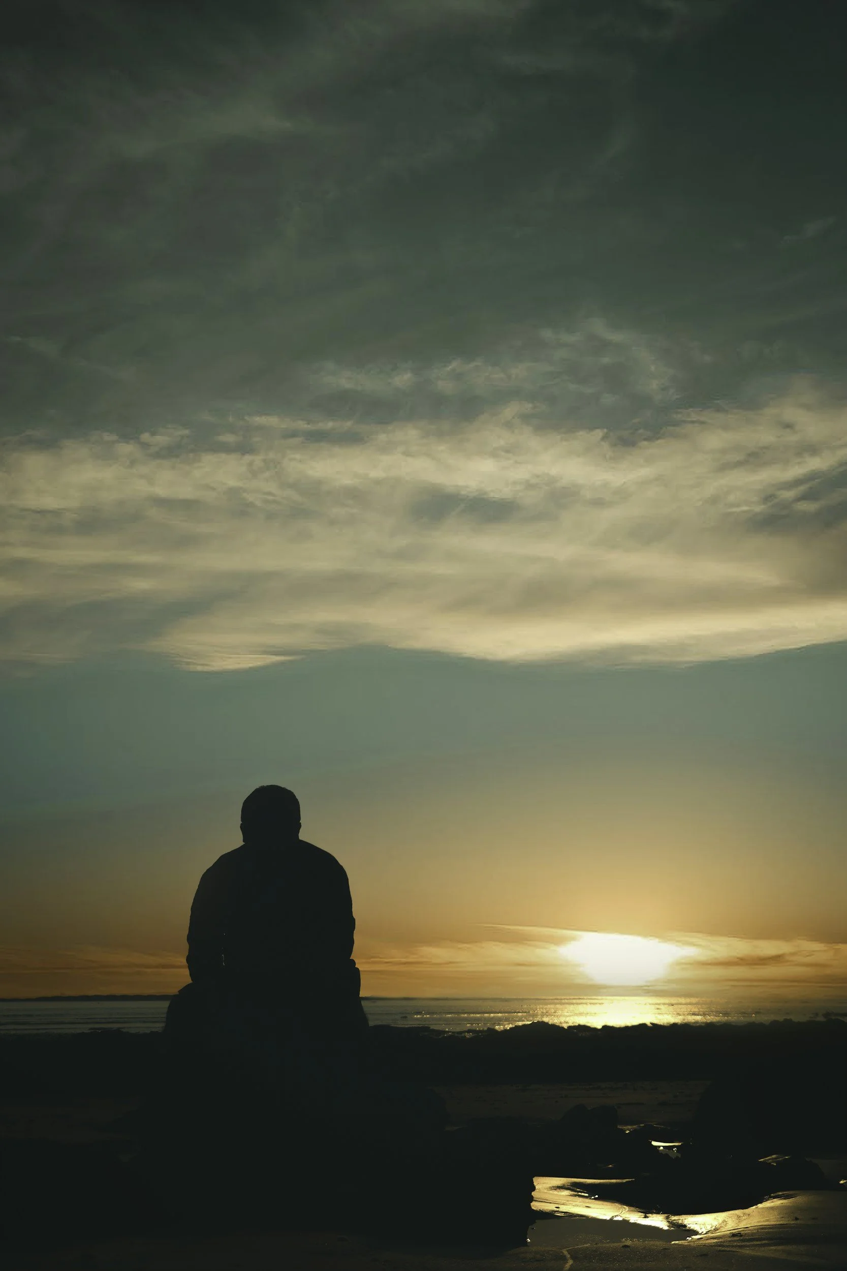 The silhouette of a person sitting by the ocean at sunset.