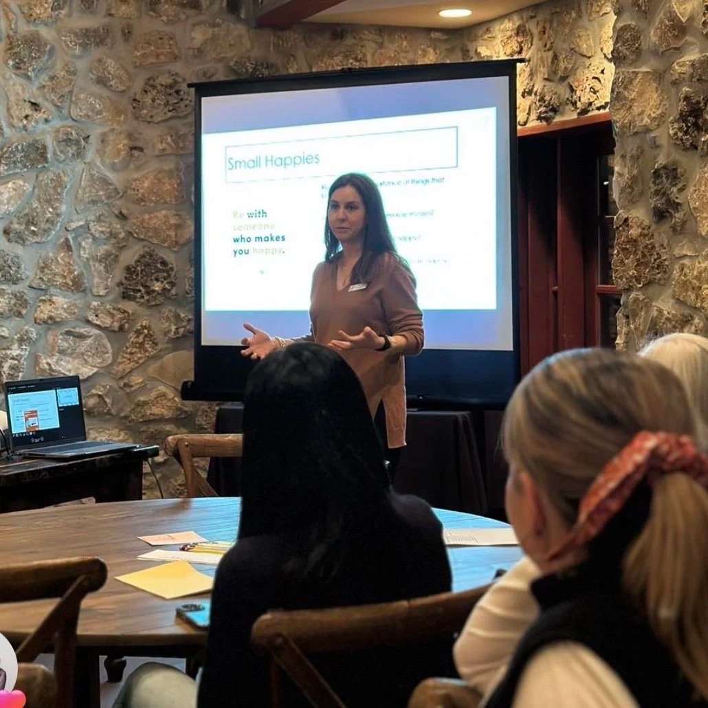 A woman giving a presentation in a room with stone walls, standing in front of a large screen displaying a slide titled 'Small Happies'. Several people are seated at a table listening, with a laptop open on the table and papers scattered around.