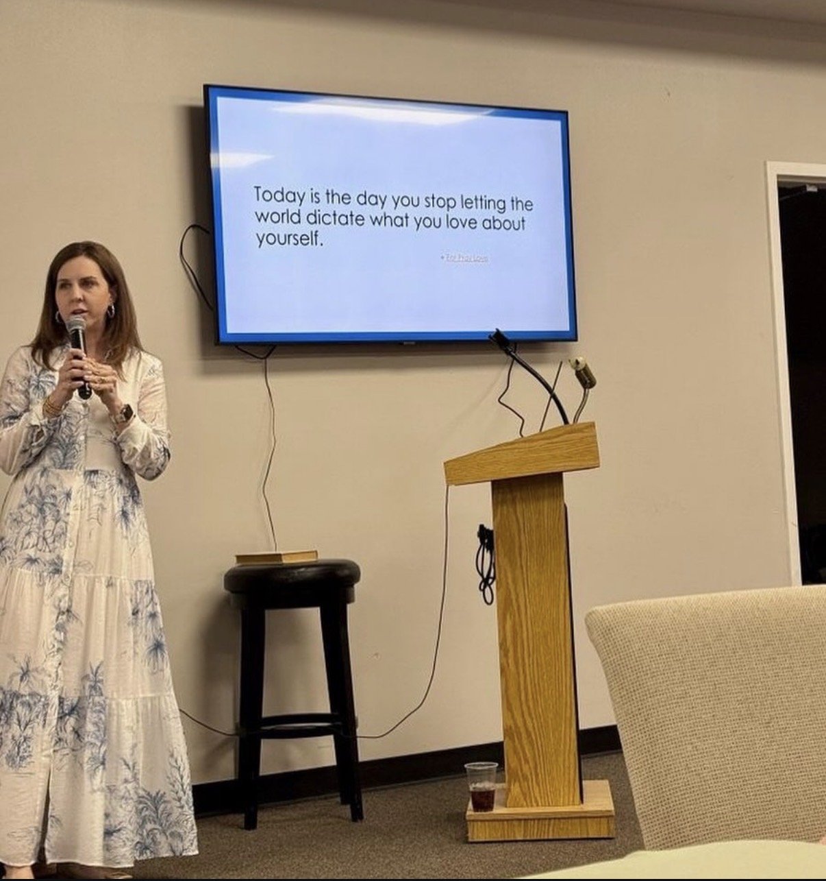 A woman speaking into a microphone in front of a large screen with a quote about self-love, in a room with beige walls and a wooden podium.