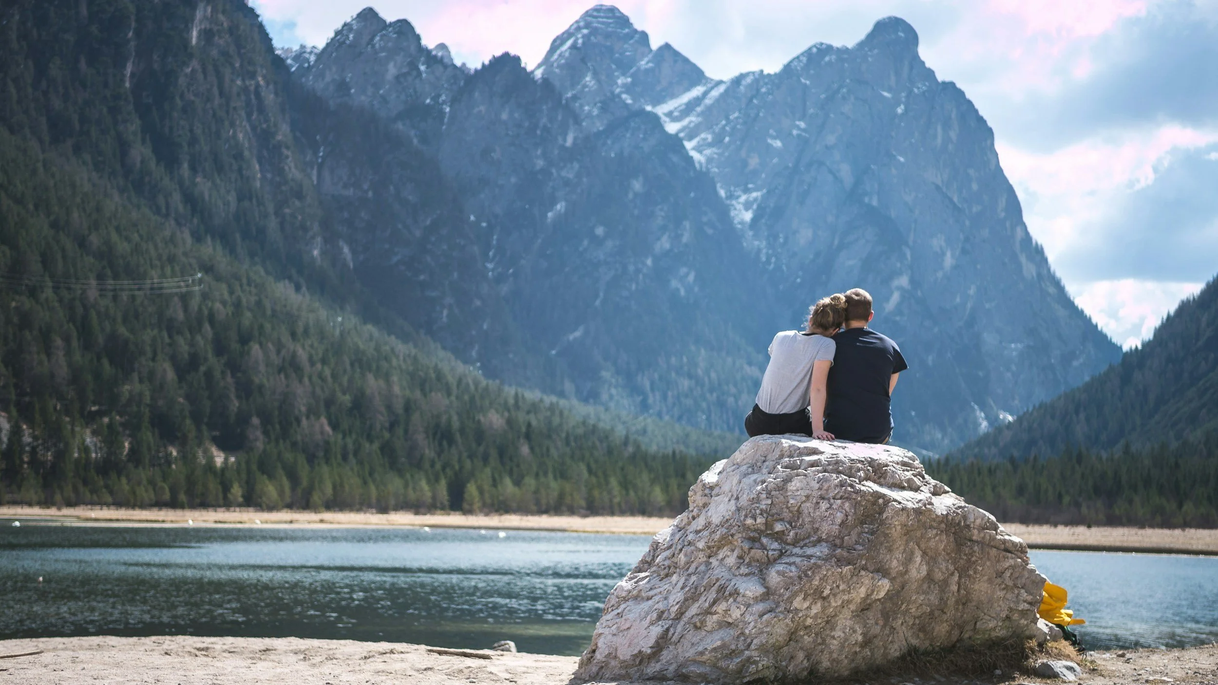 A couple sitting on a large rock by a lake, with mountains and forests in the background, enjoying a scenic view together.