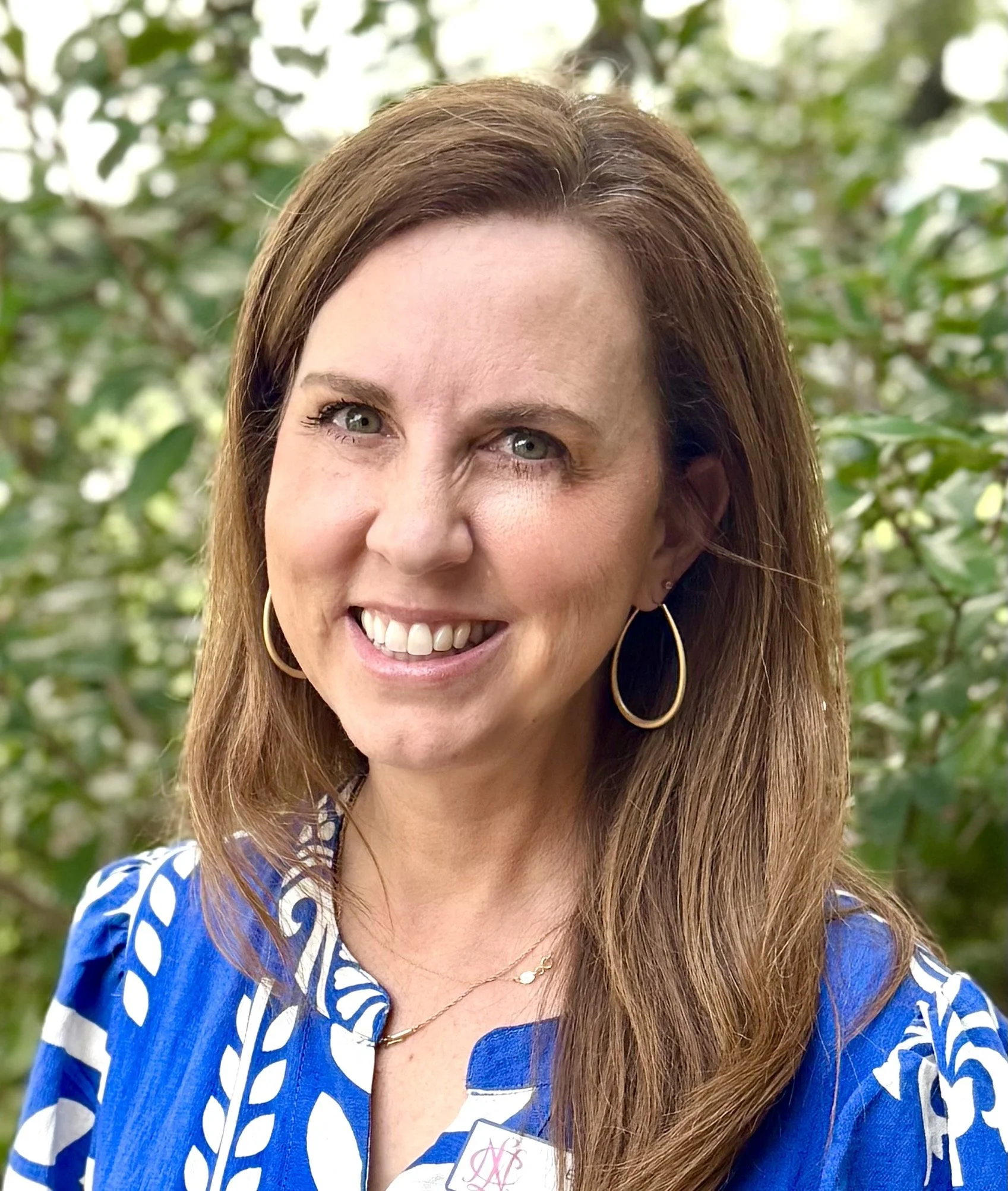 A woman with light skin, long brown hair, and green eyes smiling outdoors with green foliage in the background. She is wearing gold hoop earrings, a necklace, and a blue and white patterned dress.