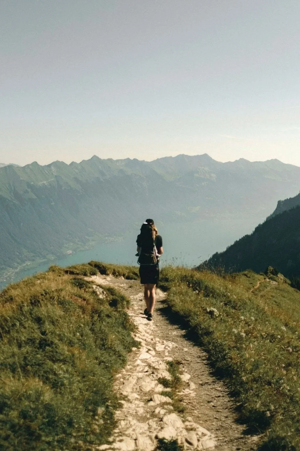 A woman hiking on a mountain trail overlooking a lake and surrounded by mountains.