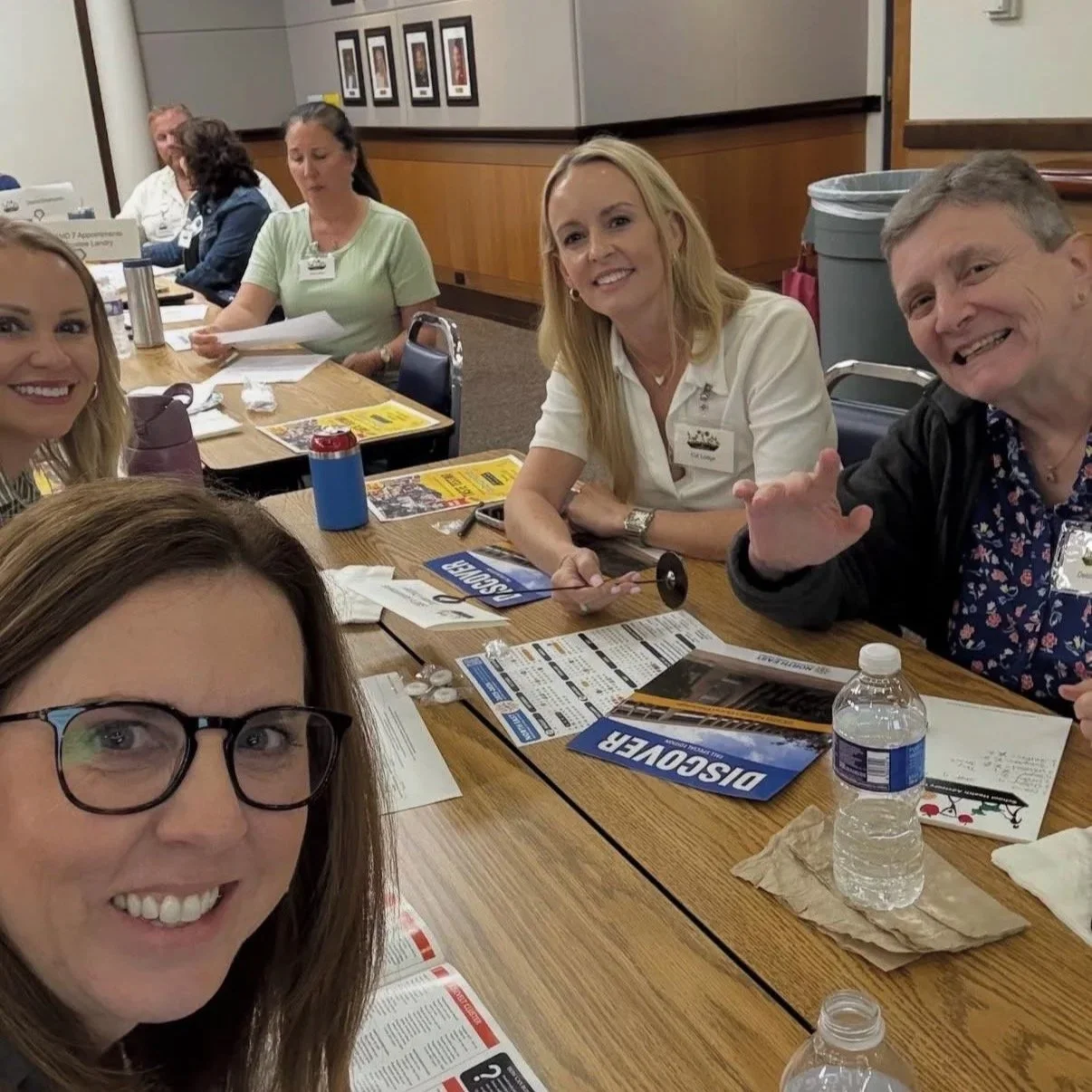 Group of people sitting around a table in a conference room, smiling, with brochures, water bottles, and papers on the table.
