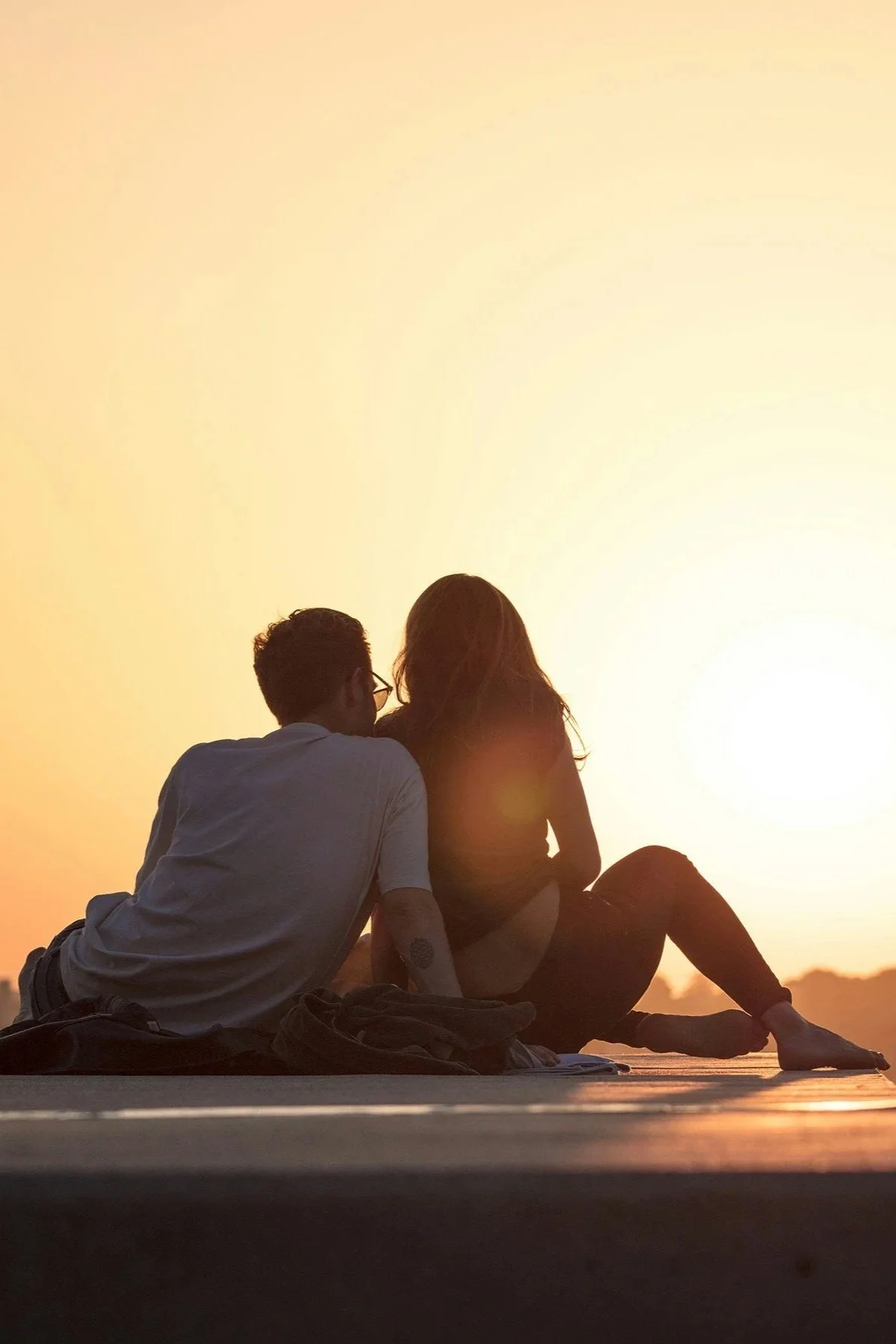 A man and a woman sitting together outdoors at sunset, with the sun setting in the background.