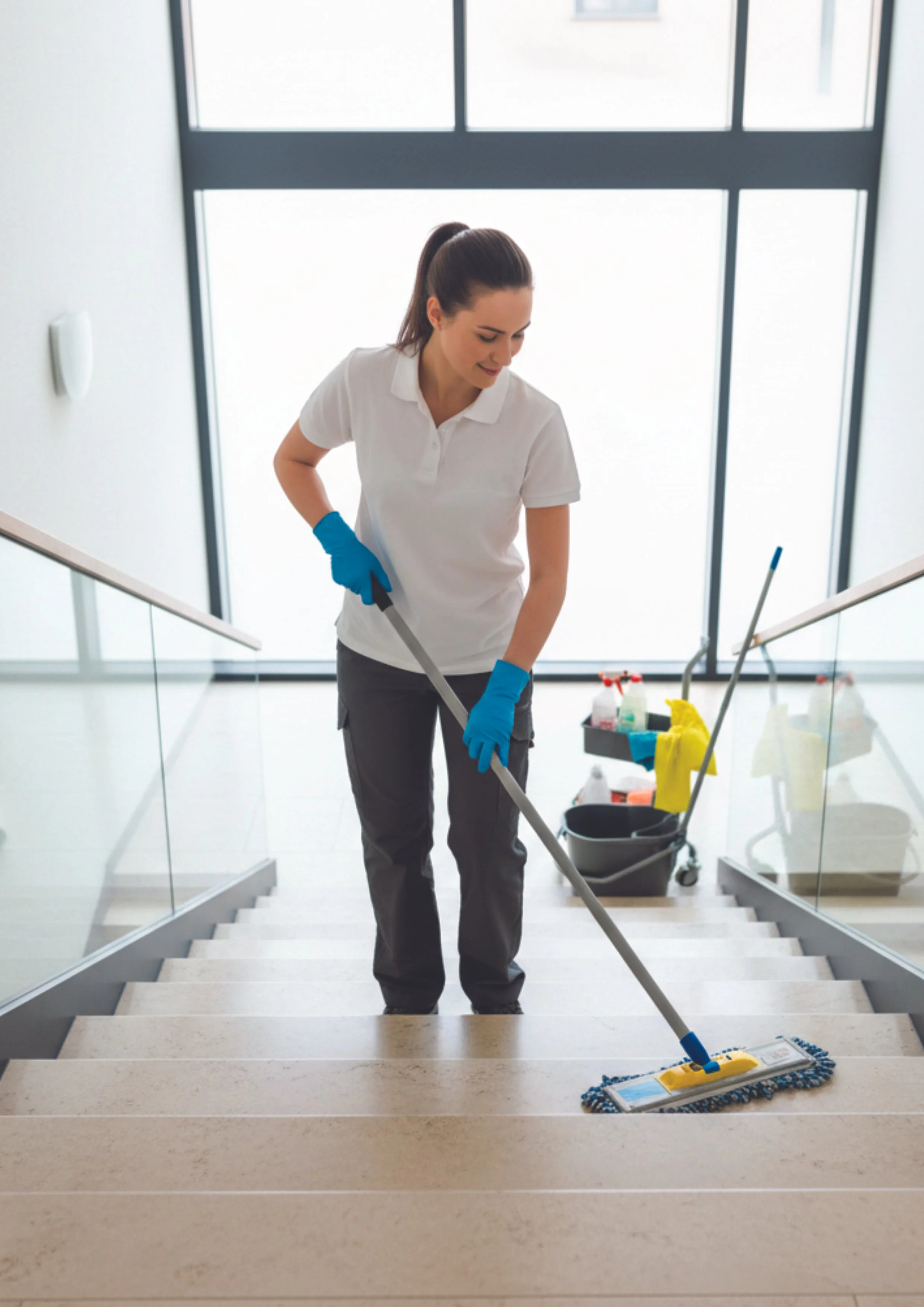 Eine Frau in weißen T-Shirt und schwarzen Hose, die auf einer Treppe mit einem Mopp beim Reinigen der Stufen arbeitet, trägt blaue Handschuhe, im Hintergrund große Fenster und Reinigungsmittel im Korb.