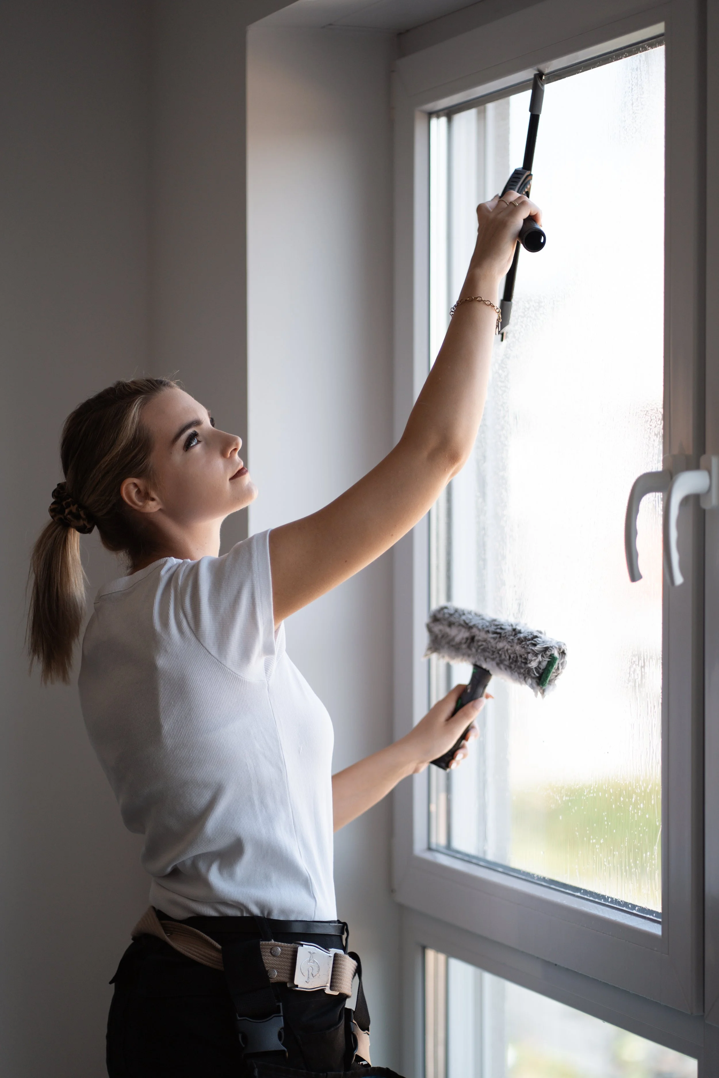 Eine junge Frau putzt ein Fenster mit einem Teleskop-Fensterwischer, trägt einen weißen T-Shirt und eine schwarze Hose mit einem Gürtel.