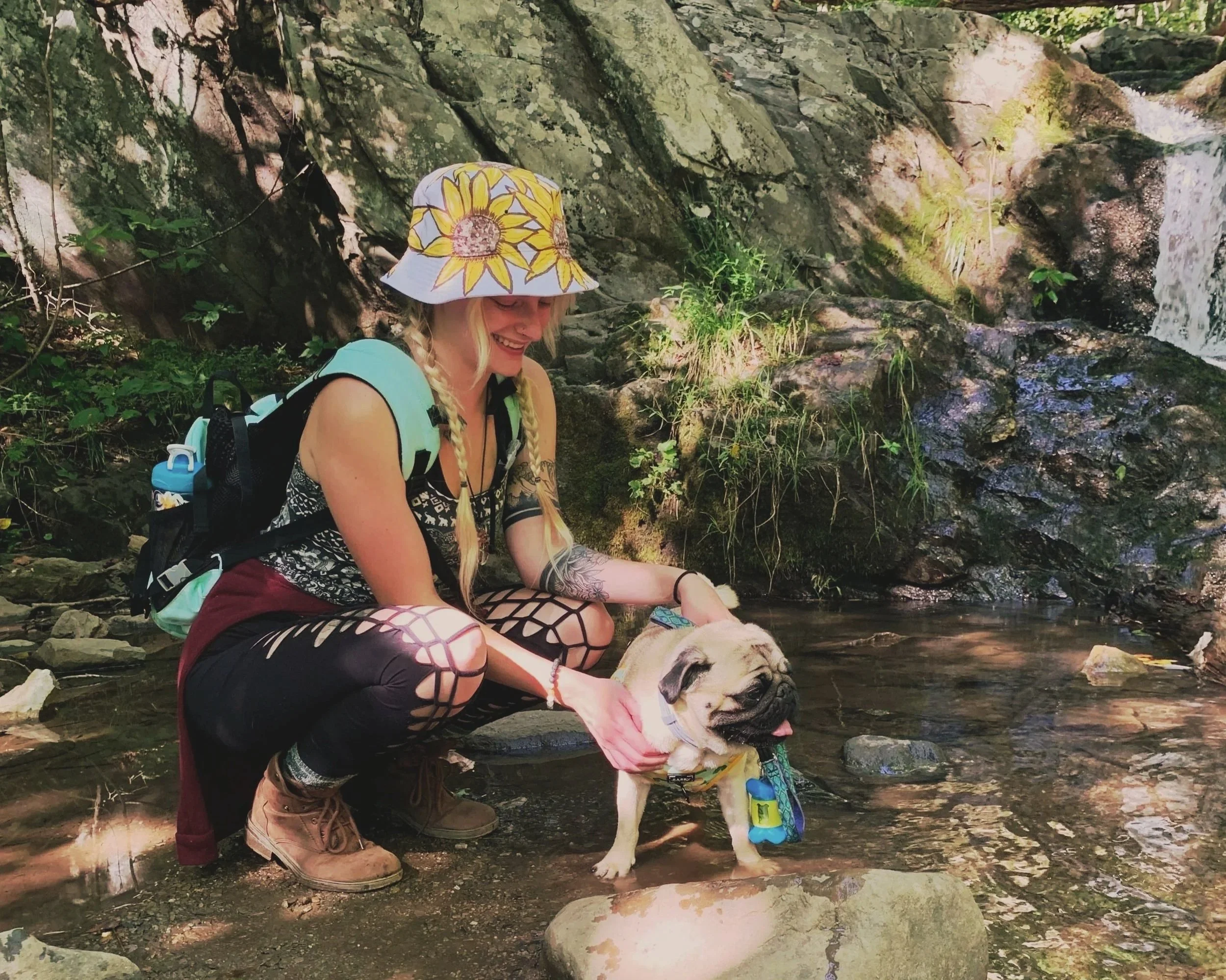 A woman with blonde hair, wearing a sun hat with sunflower patterns, a patterned top, and fishnet stockings, is smiling and sitting by a river with a dog. The dog, a pug, is wearing a colorful outfit and is standing in the water. There is a rocky, green forest background with sunlight filtering through the trees.