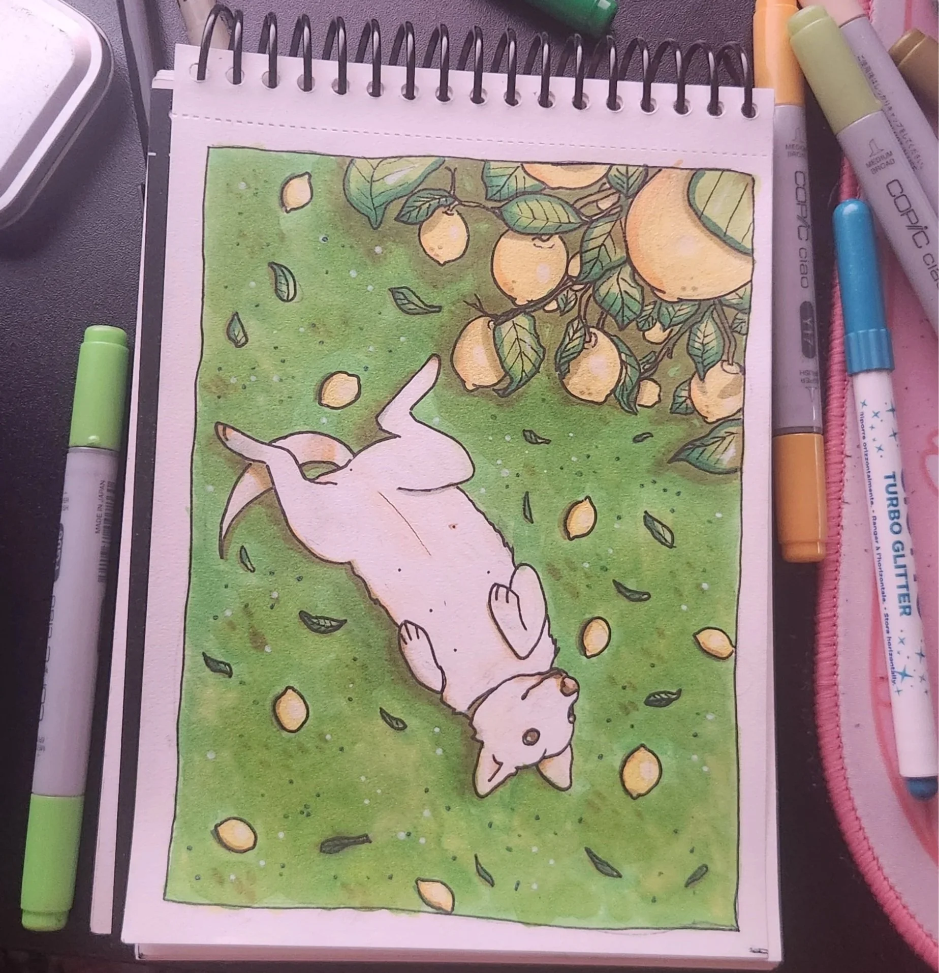 A watercolor painting of a lemon tree with yellow lemons, green leaves, and some falling lemons on a green background. There are various markers and pens around the drawing.
