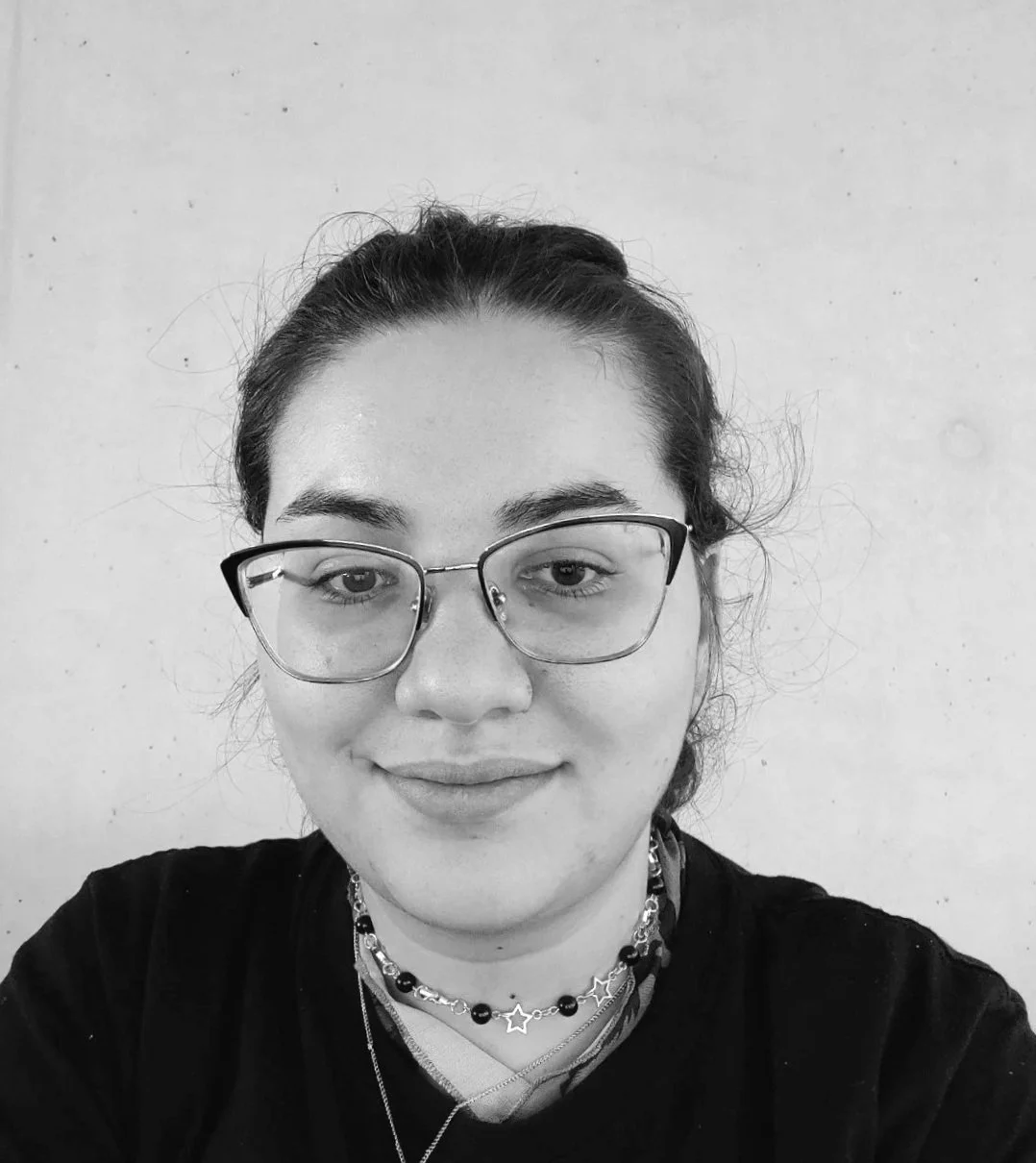 Black and white photo of a young woman with glasses, dark hair tied back, smiling slightly, wearing layered necklaces, black shirt, against a plain light background.