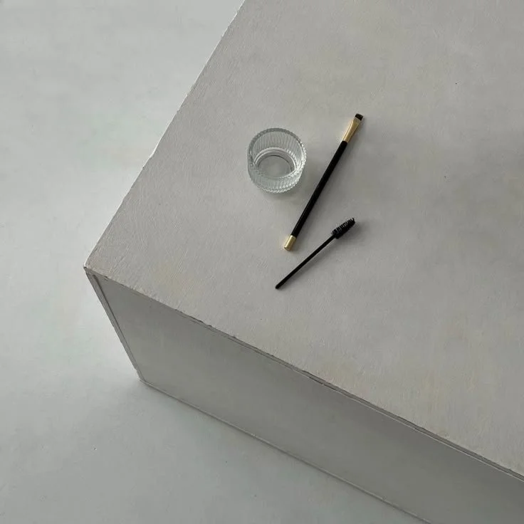 A glass of water, an eyelash brush, and a makeup brush on a white surface.