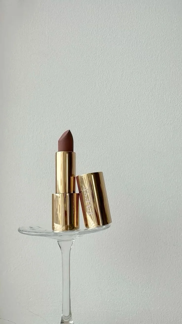A lipstick with a gold casing standing upright on a glass stand, with the cap leaning against it, against a plain light grey background.