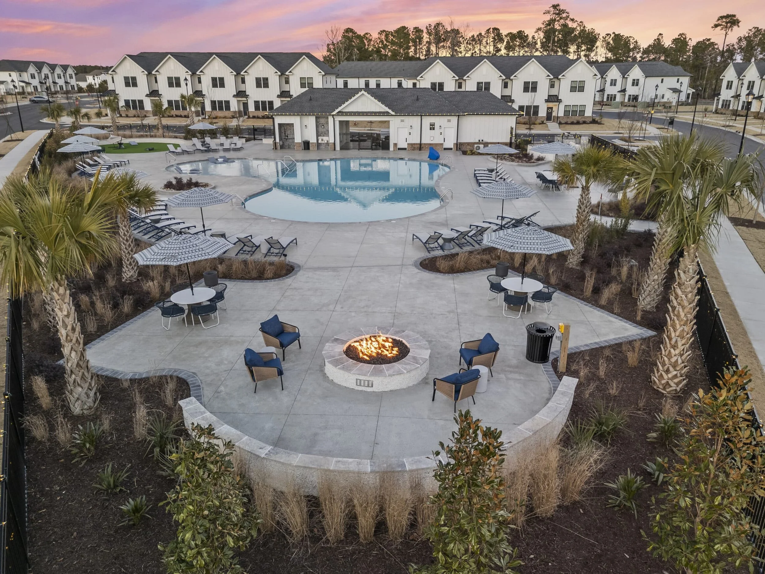 Townes at Blake Farm A community swimming pool area with lounge chairs, umbrellas, and a fire pit, surrounded by palm trees and townhouses under a colorful sunset sky.