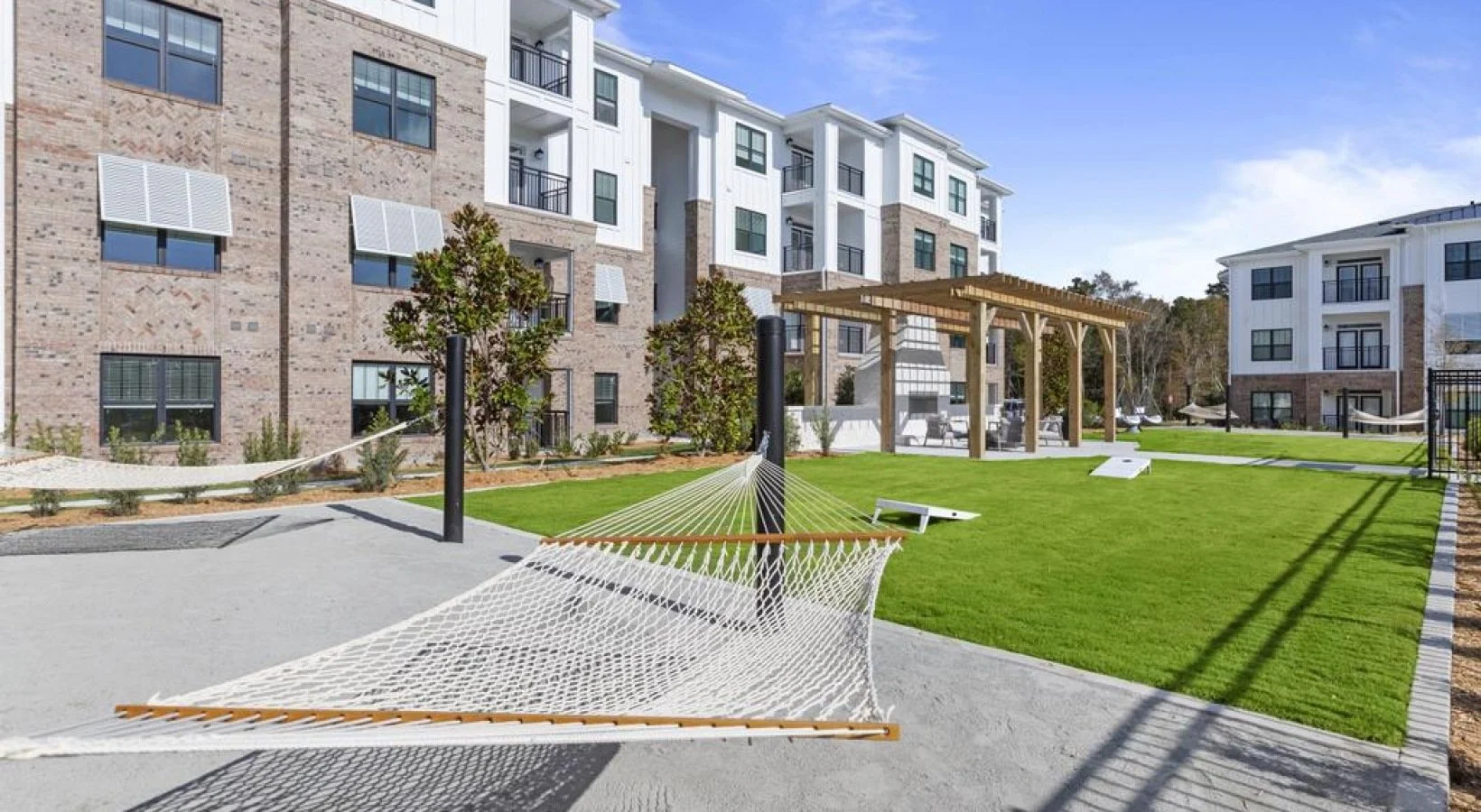 Reserve at Blake Farm Outdoor recreational area in an apartment complex with a hammock, artificial grass, a wooden pergola, parking spots, and modern multi-story apartment buildings in the background.
