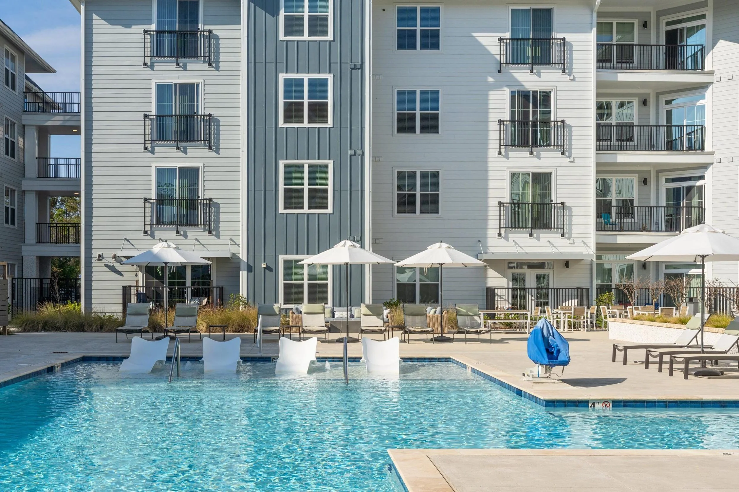 Oak Grove at Blake Farm View of a residential apartment complex with balconies, white and gray exterior walls, and a swimming pool in the foreground. Lounge chairs and umbrellas are set up near the pool.