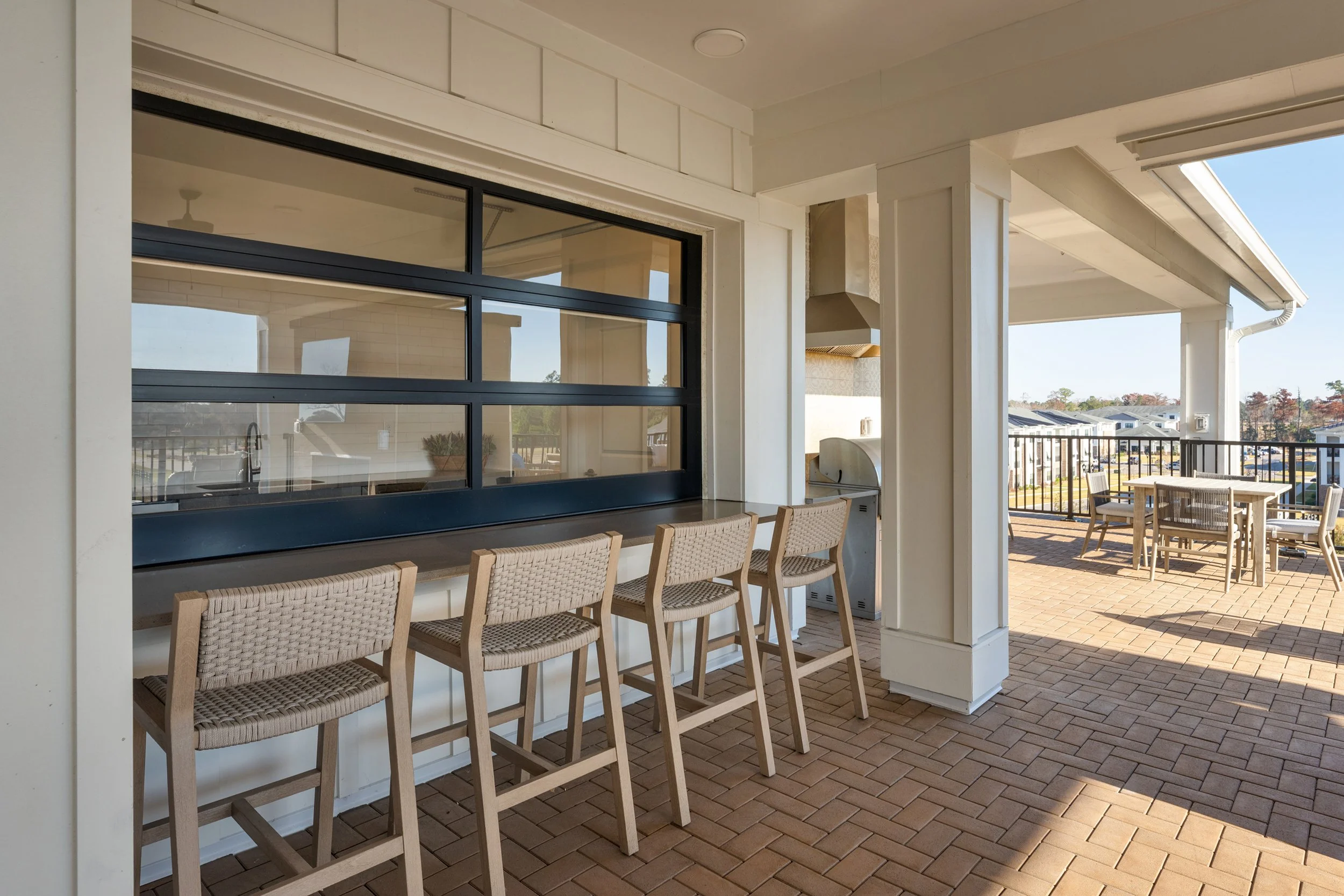 Oak Grove at Blake Farm Outdoor balcony area with a row of chairs, a high counter with bar stools, and a dining table with chairs, overlooking neighboring houses and trees.
