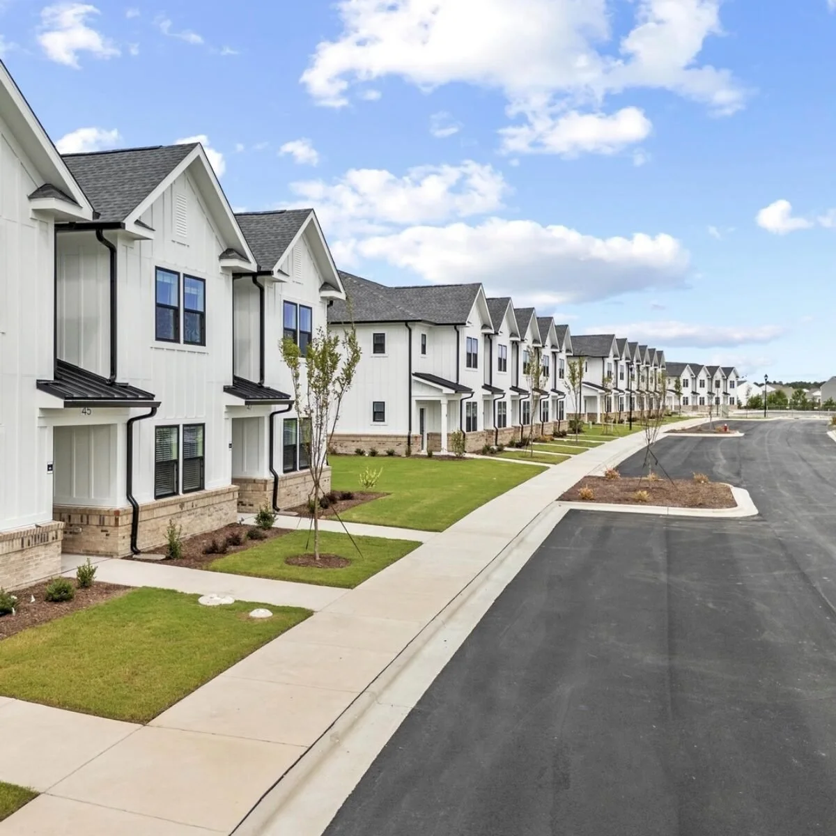 Townes at Blake Farm Row of modern white townhouses with small front yards and newly planted trees, along a paved street on a partly cloudy day.