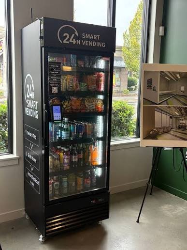 A vending machine labeled "24H Smart Vending" stocked with snacks and beverages, located indoors next to a window, with a digital payment terminal attached.