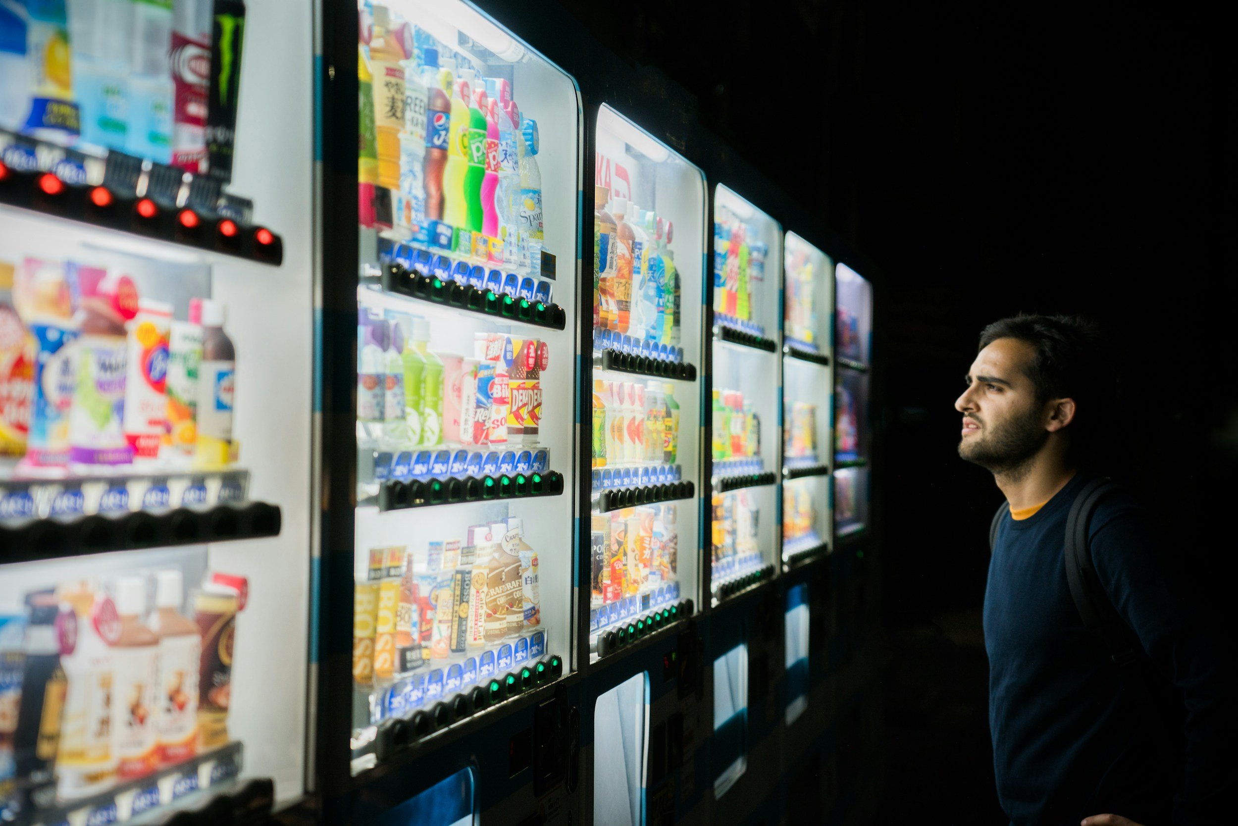 A man shopping at a vending machine filled with various bottled beverages, located in a dark area.