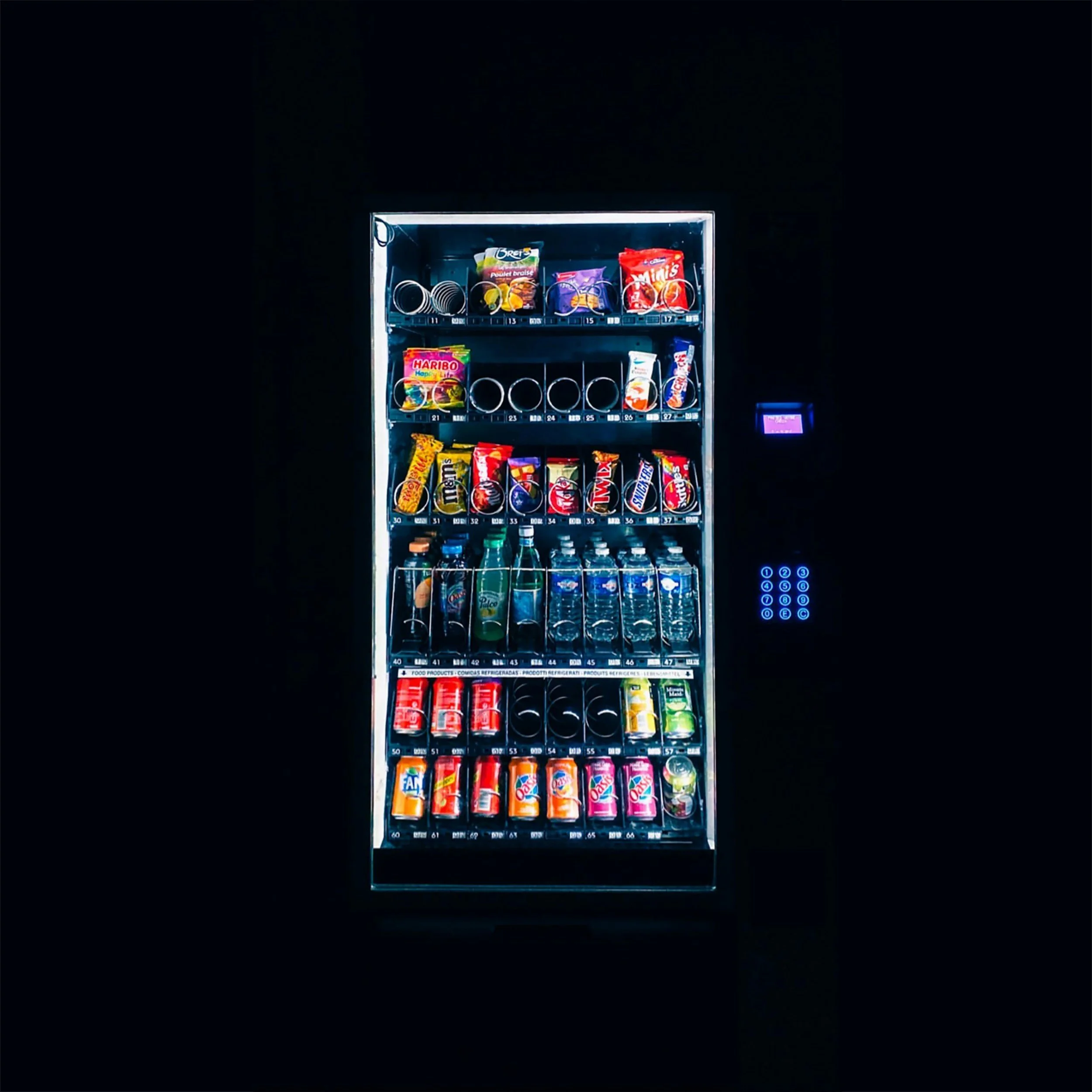 A vending machine illuminated in a dark room, stocked with snacks, chips, candies, and bottled drinks.