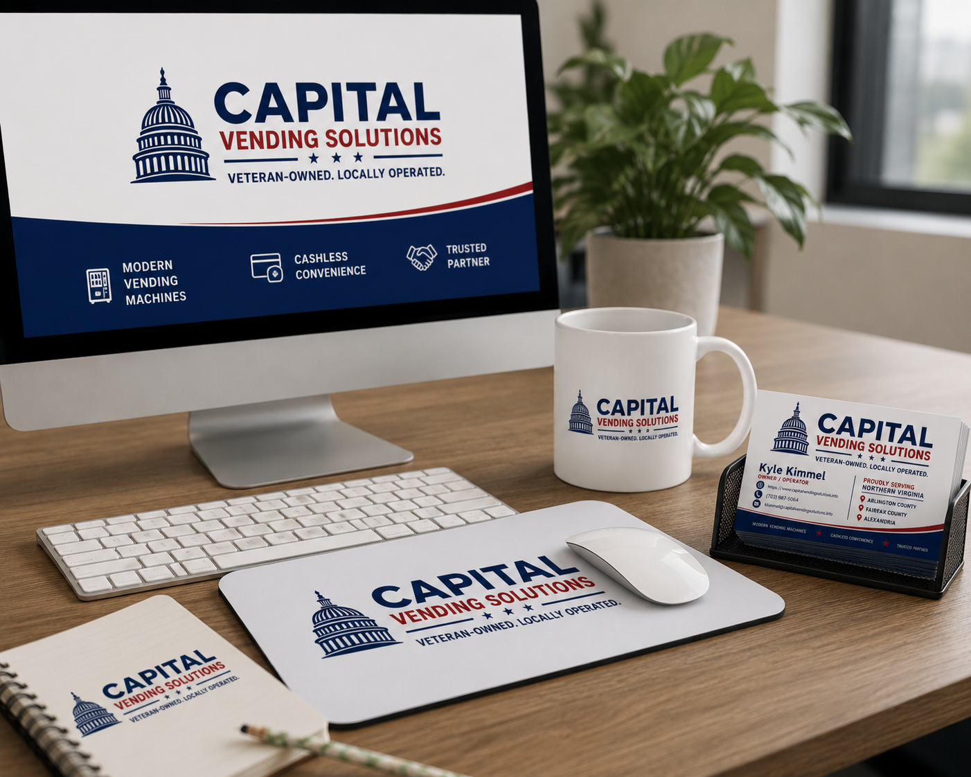 Desk setup featuring branding materials for Capital Vending Solutions, including a computer monitor, branded mug, company business card holder with business cards, mouse, mouse pad, notepad, and a potted plant in the background.