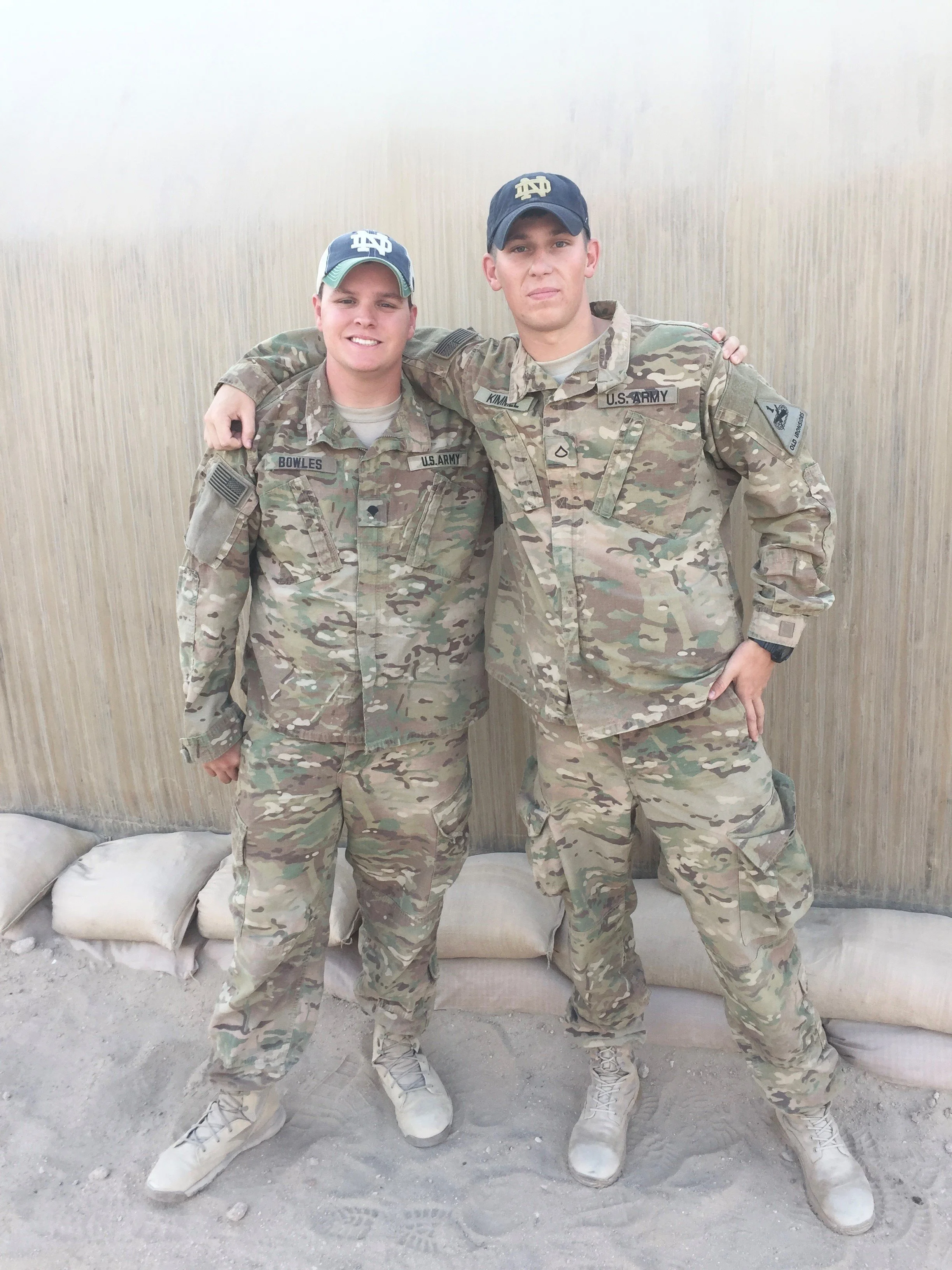 Two soldiers in camouflage uniforms pose together, with one soldier's arm around the other's shoulder, standing in front of a beige wall.
