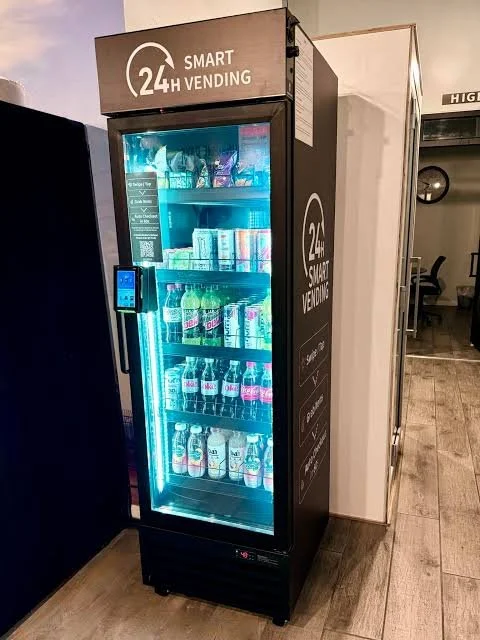 A black vending machine with a glass door illuminated by blue lights, stocked with beverages including soda, water, and juice. It has a sign on top that reads '24/7 Smart Vending'.