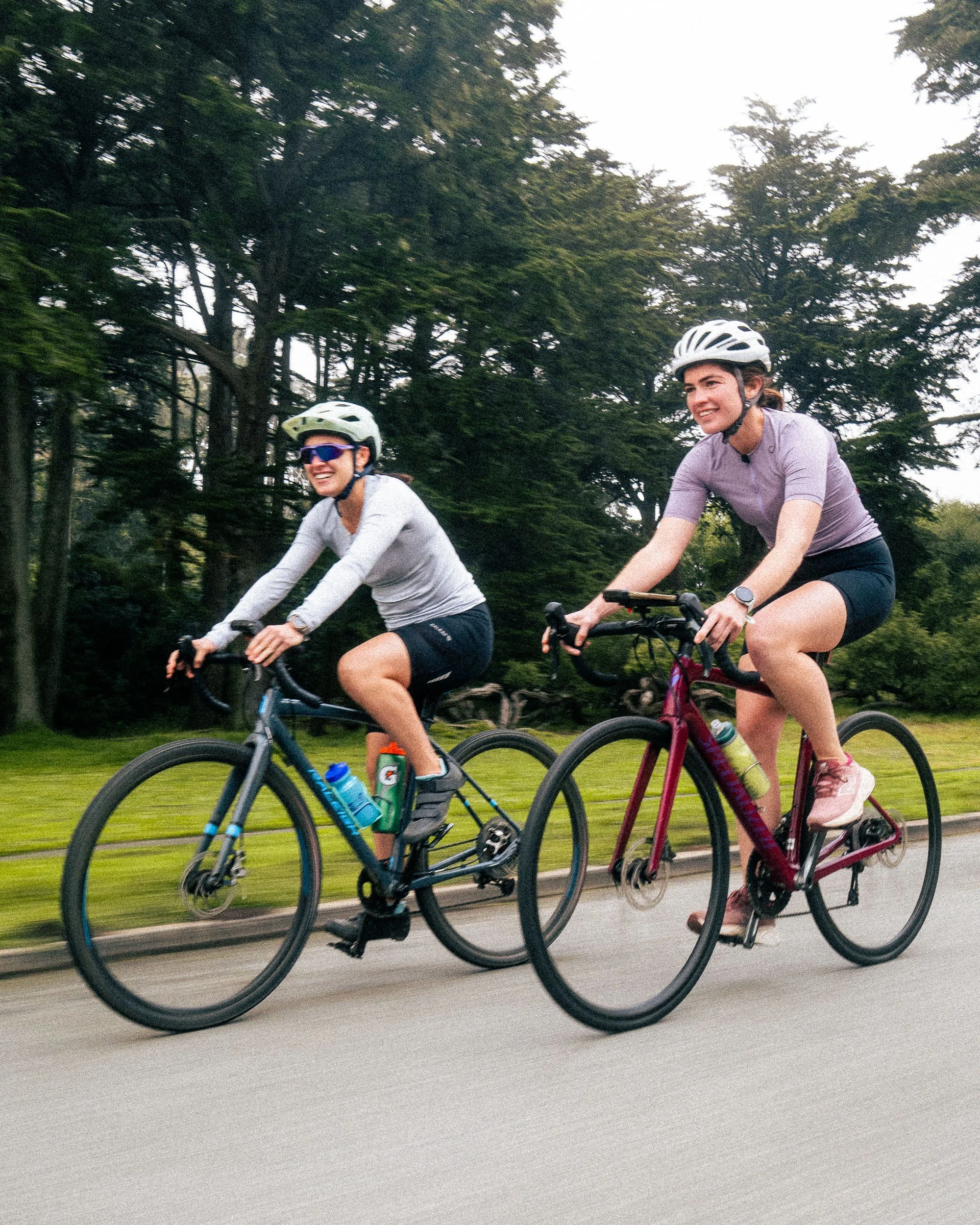 Two women riding bicycles outdoors on a paved trail with green grass and trees in the background, wearing helmets and athletic clothing, smiling and enjoying a daytime ride.