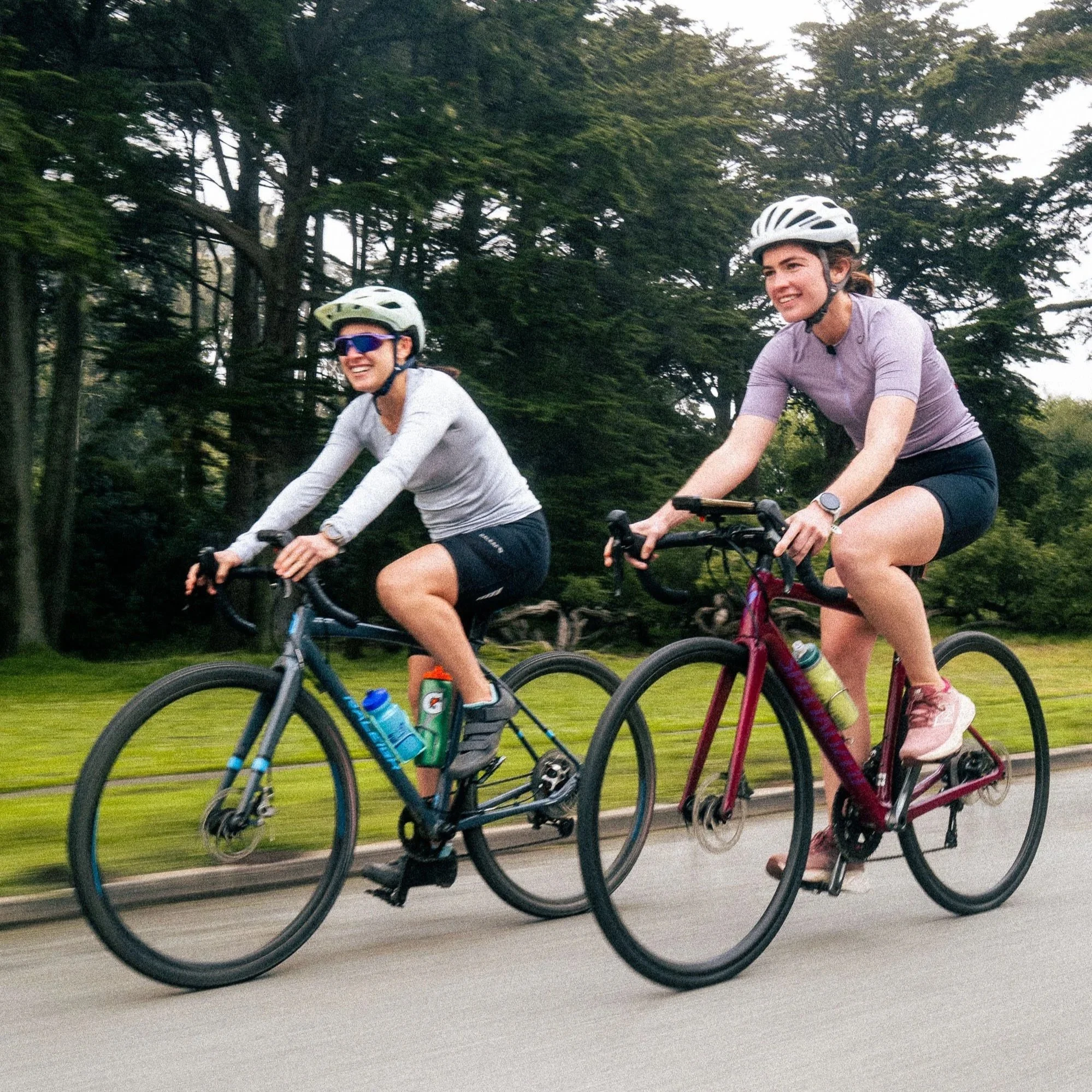 Two women riding bikes outdoors on a paved trail, both wearing helmets and casual athletic clothing, with trees and greenery in the background.
