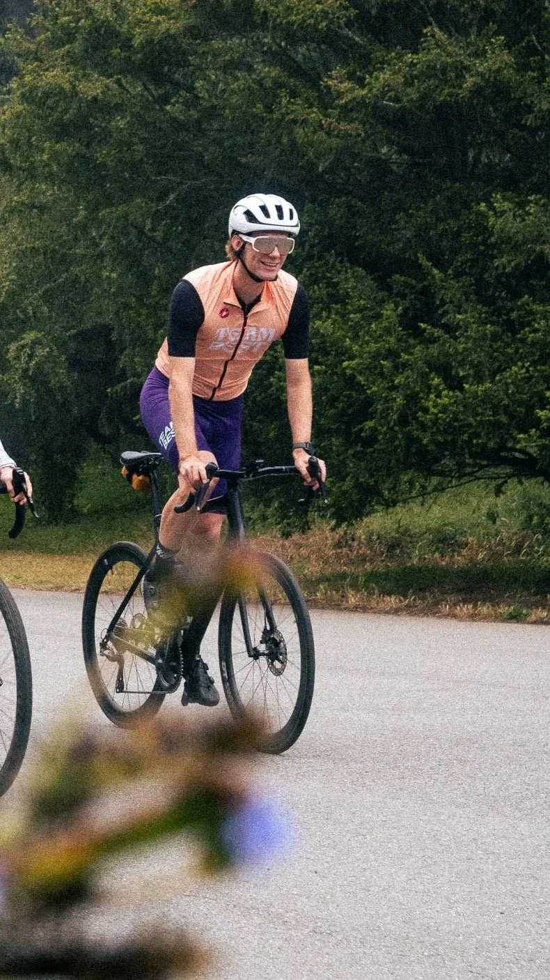 A person riding a black bicycle on a paved road, wearing a white helmet, orange and black cycling jersey, purple shorts, and sunglasses, with green trees in the background.