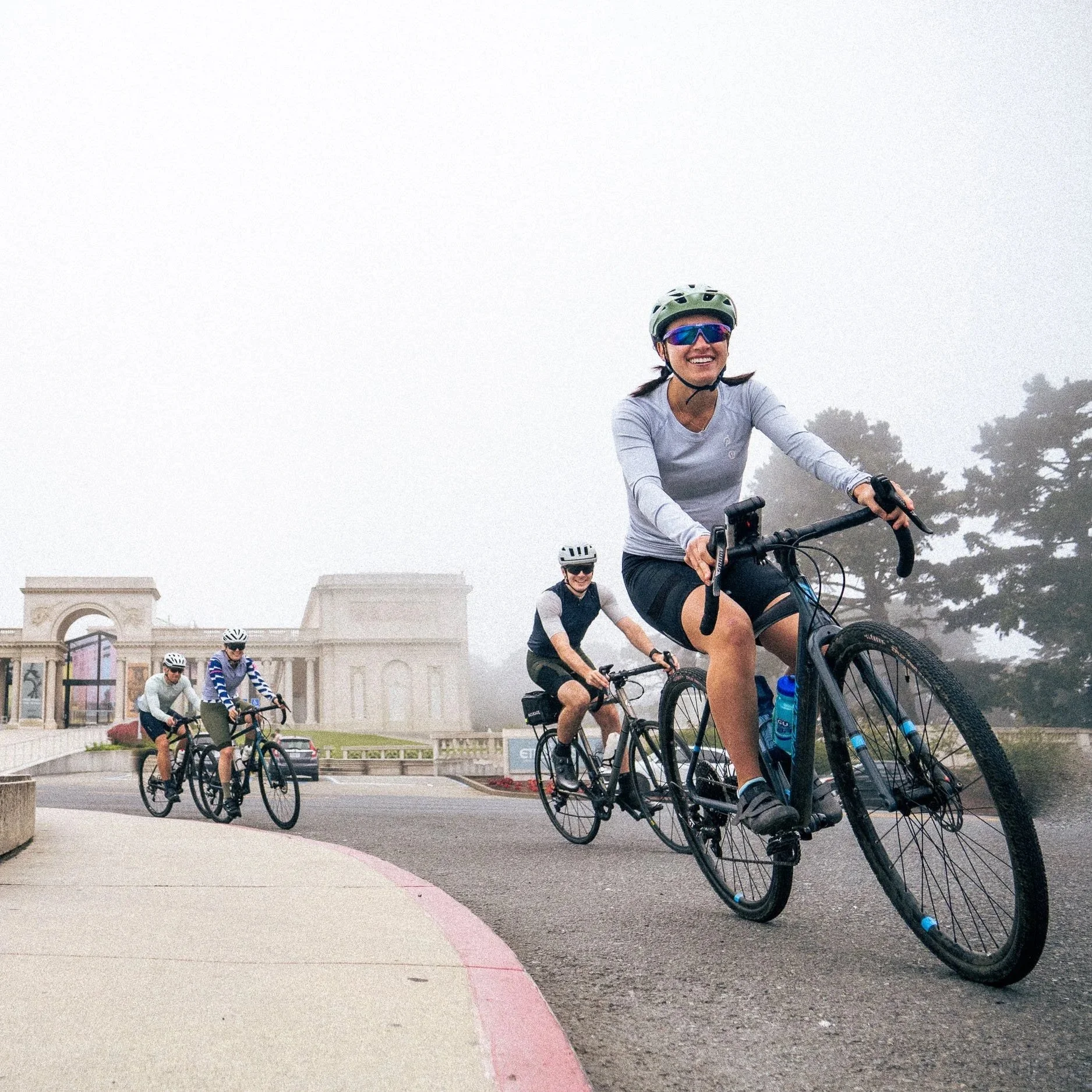 A group of people riding bikes on a city street in foggy weather, with a landmark building and trees in the background.